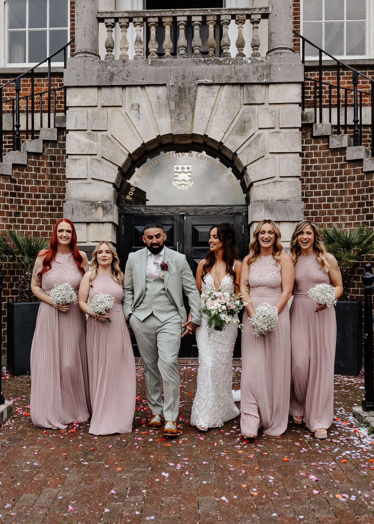Wedding party in dusty rose dresses and gray suit poses together with confetti scattered on ground near brick building.