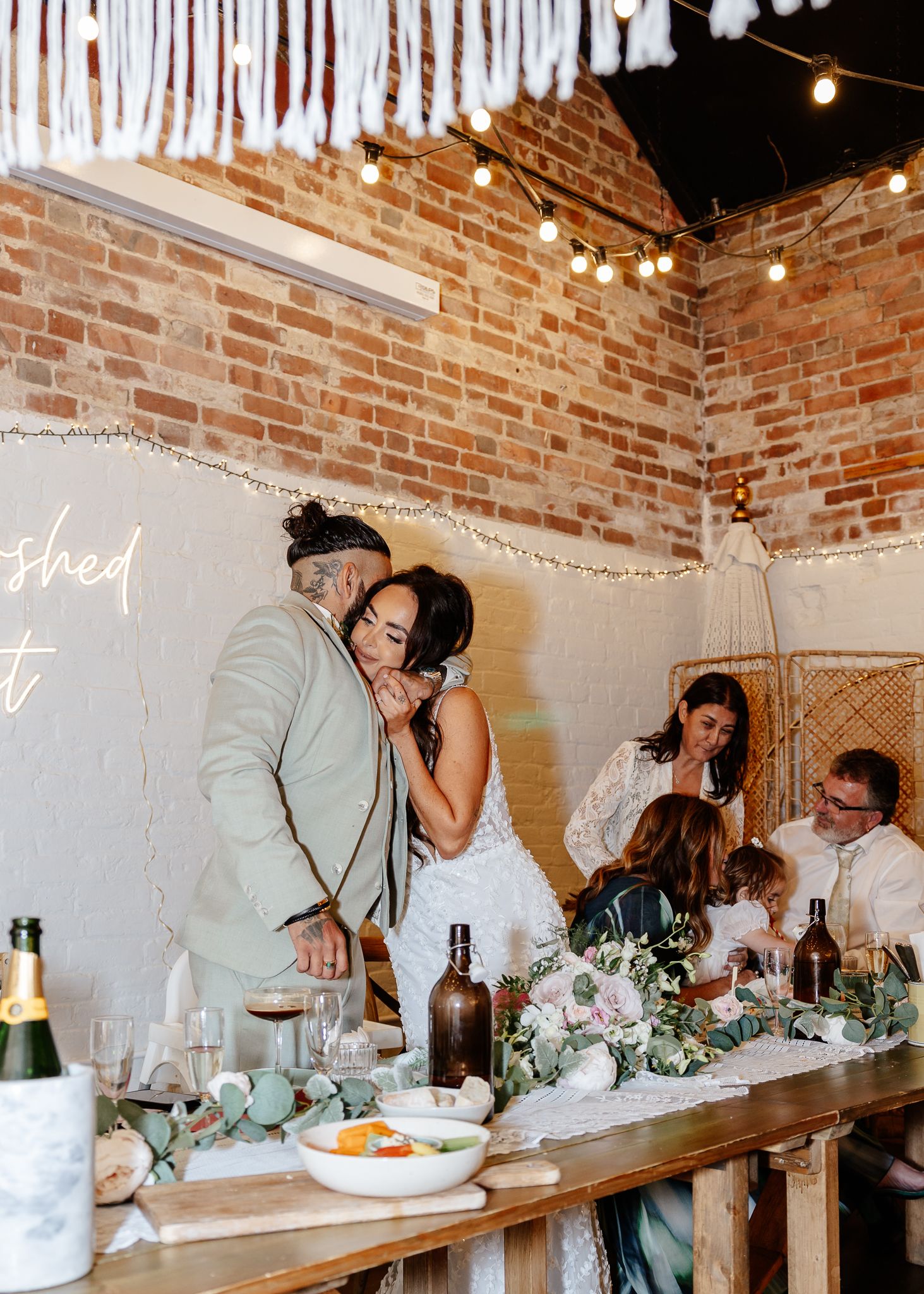 Wedding guests seated at a rustic long table with string lights and exposed brick walls in an intimate reception venue.