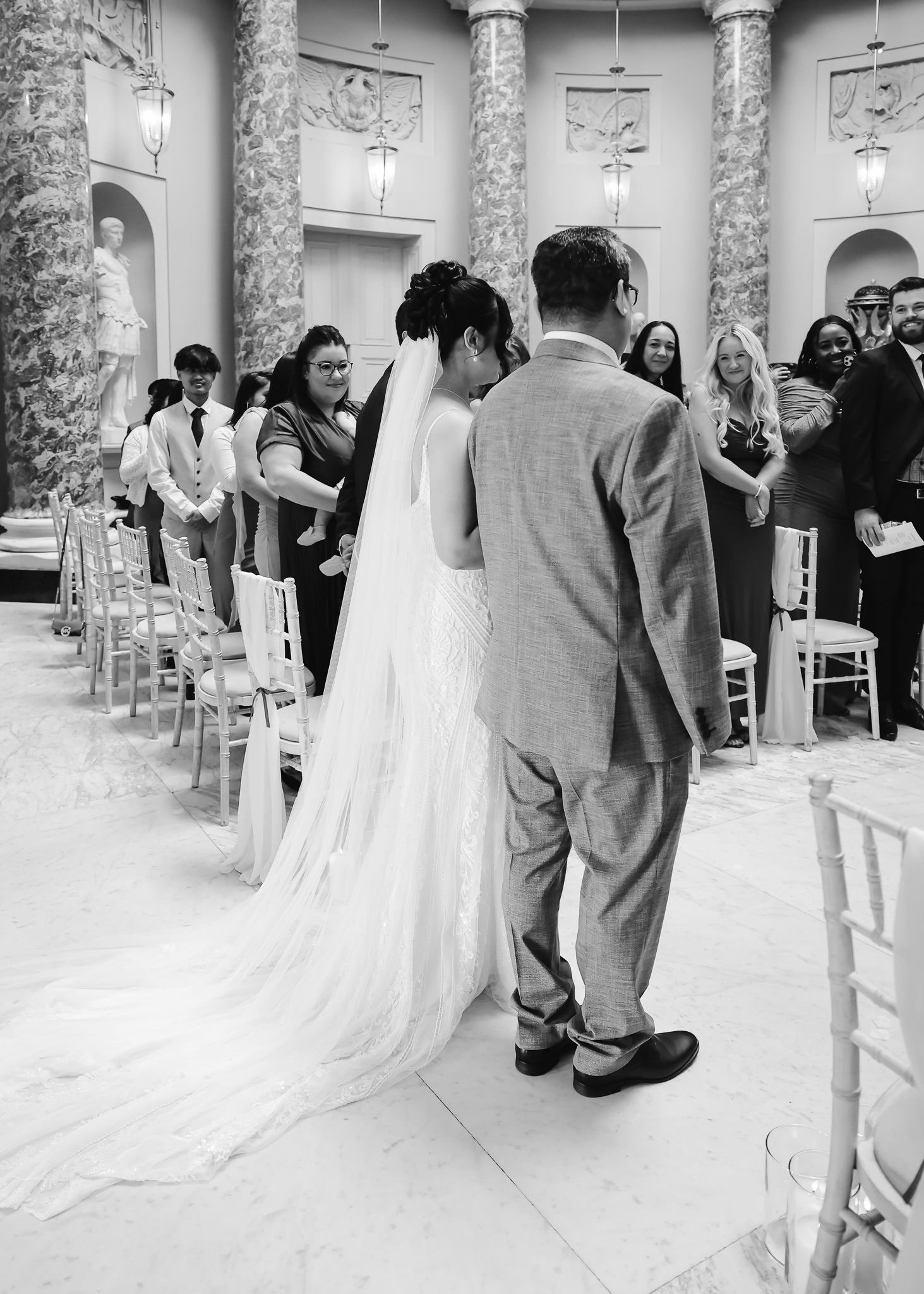 Wedding ceremony procession in an elegant indoor venue with marble columns and white chairs.