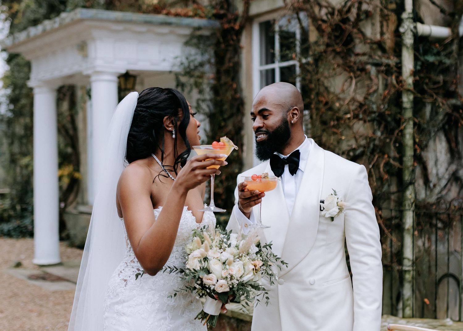 Newlywed couple shares romantic moments in a garden setting with white pillars and floral arrangements.