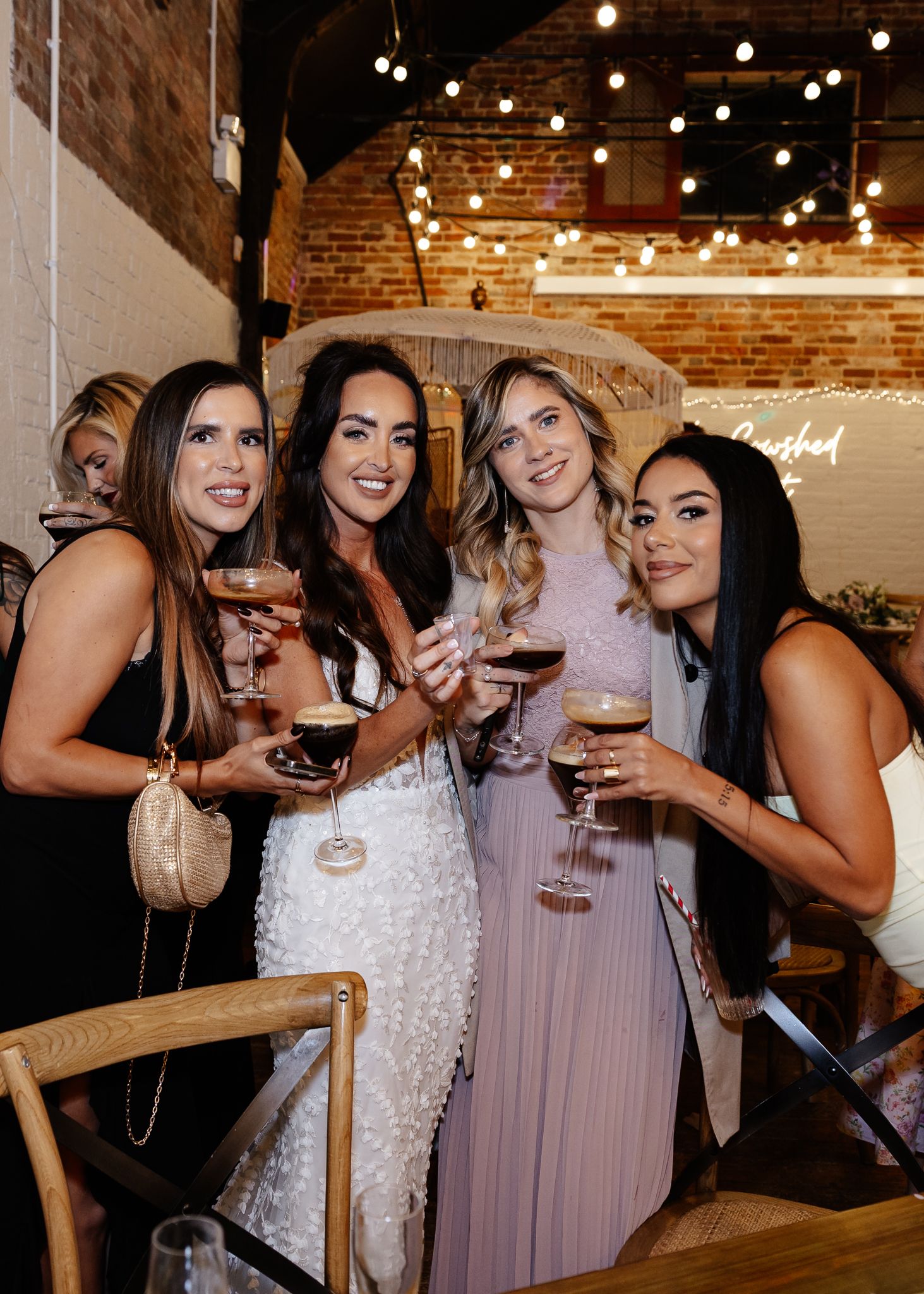Wedding party enjoying drinks and laughter at a rustic brick venue with string lights overhead.