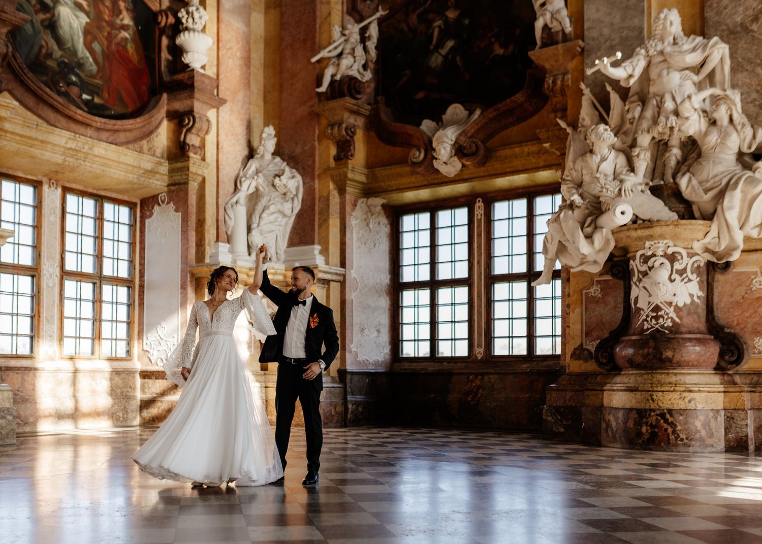 Panoramic view of an ornate baroque ballroom with marble floors, statues and sunlit windows in a historic palace.