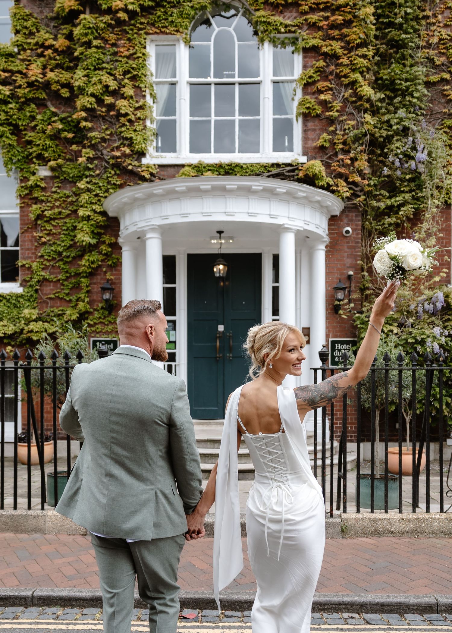 A bride and groom hold hands outside an ivy-covered brick wedding venue, bride raising bouquet.
