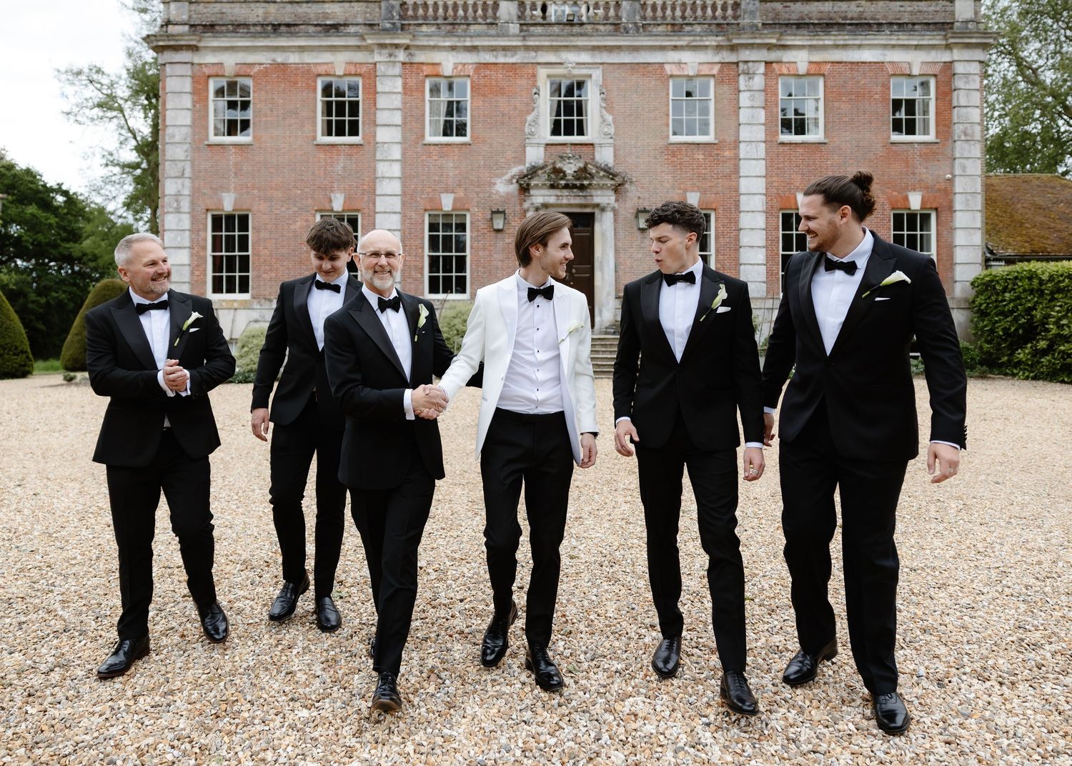 Groom in white tuxedo stands with six groomsmen in black tuxedos outside a brick manor house.