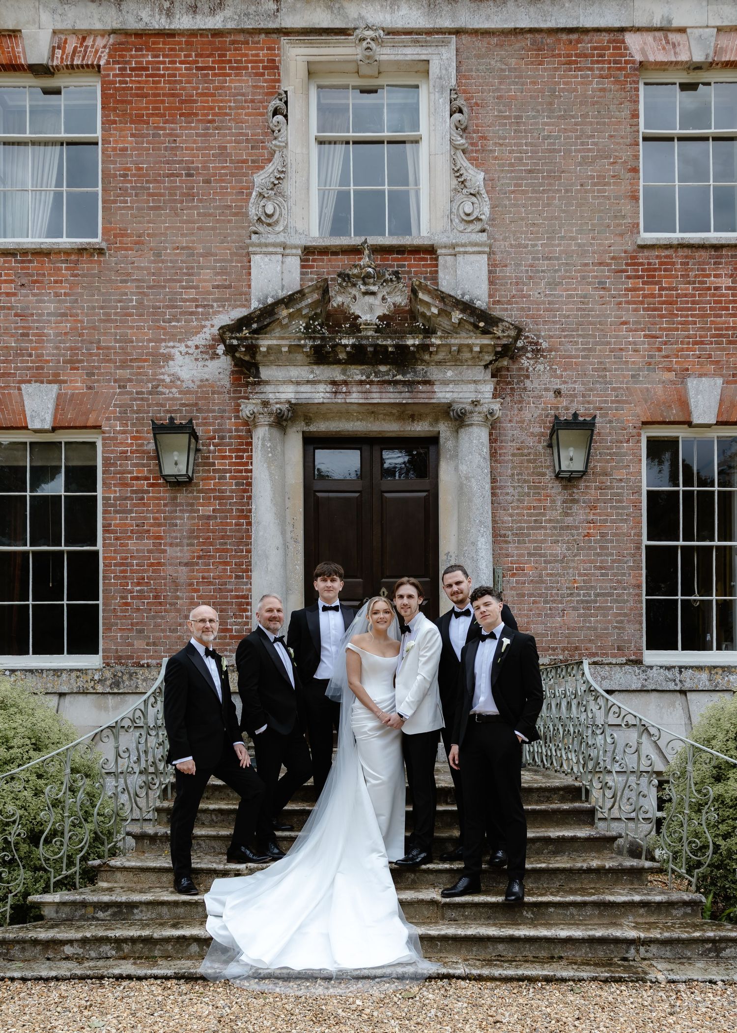Wedding party poses on grand brick manor steps, bride in white gown with train, groom and groomsmen in black tuxedos.