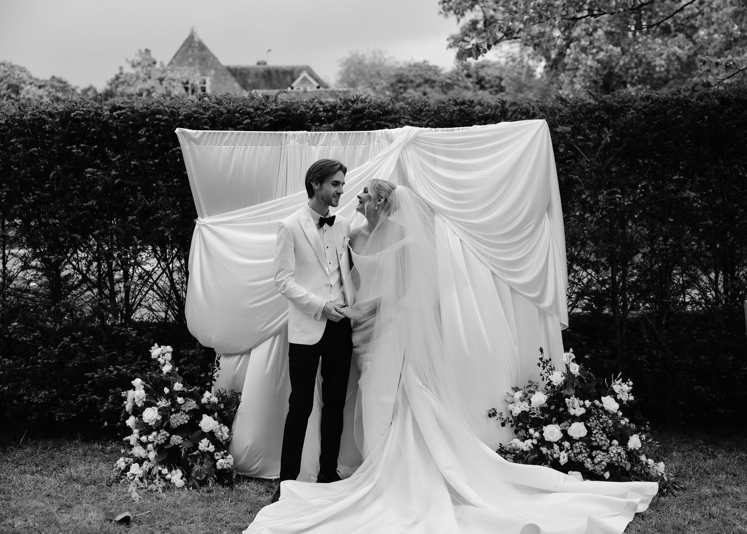 Black and white wedding photo of couple embracing before draped white backdrop with floral arrangements outdoors.