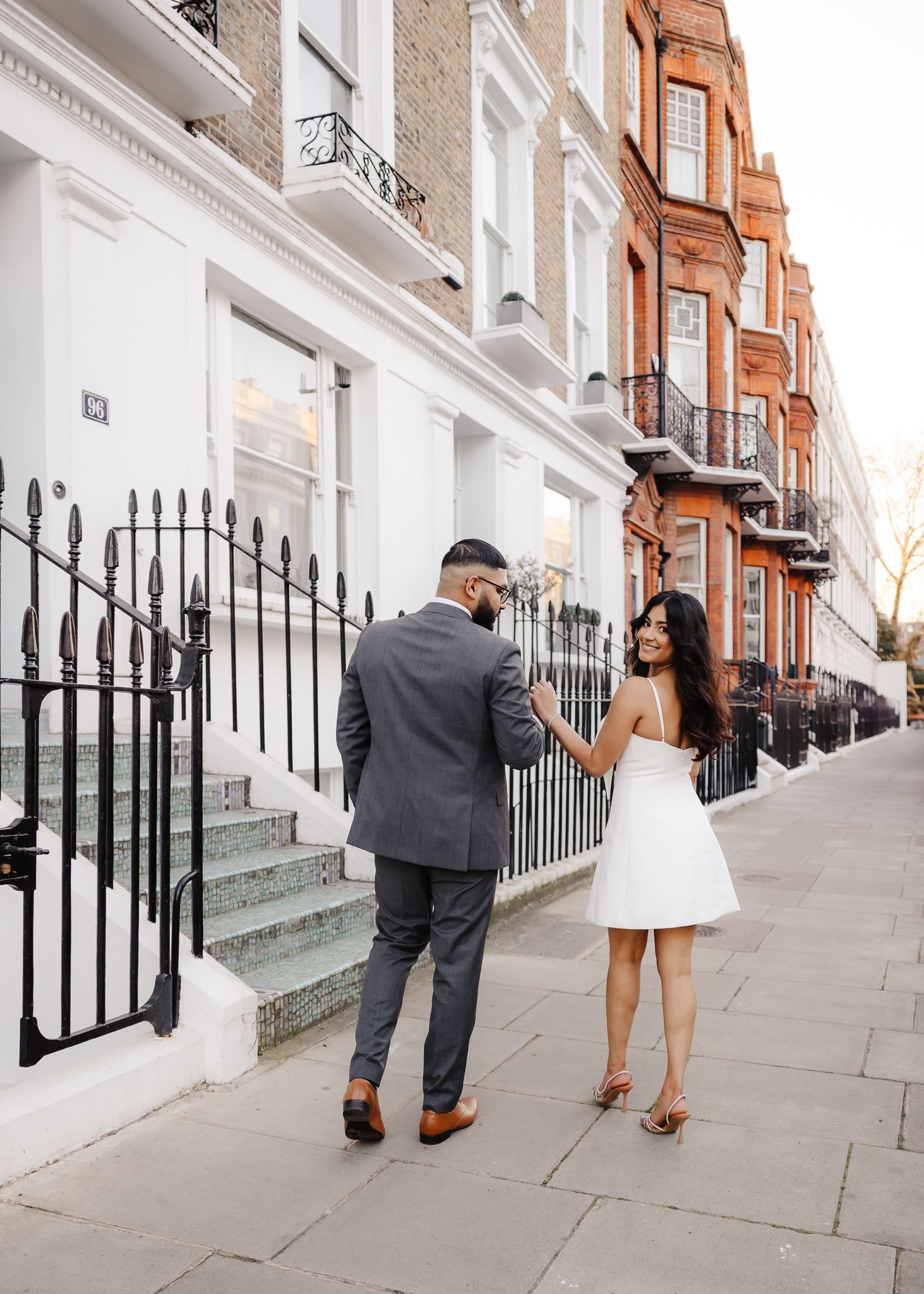 Couple walking on a London street, woman in white dress, man in grey suit, beside red brick townhouses.