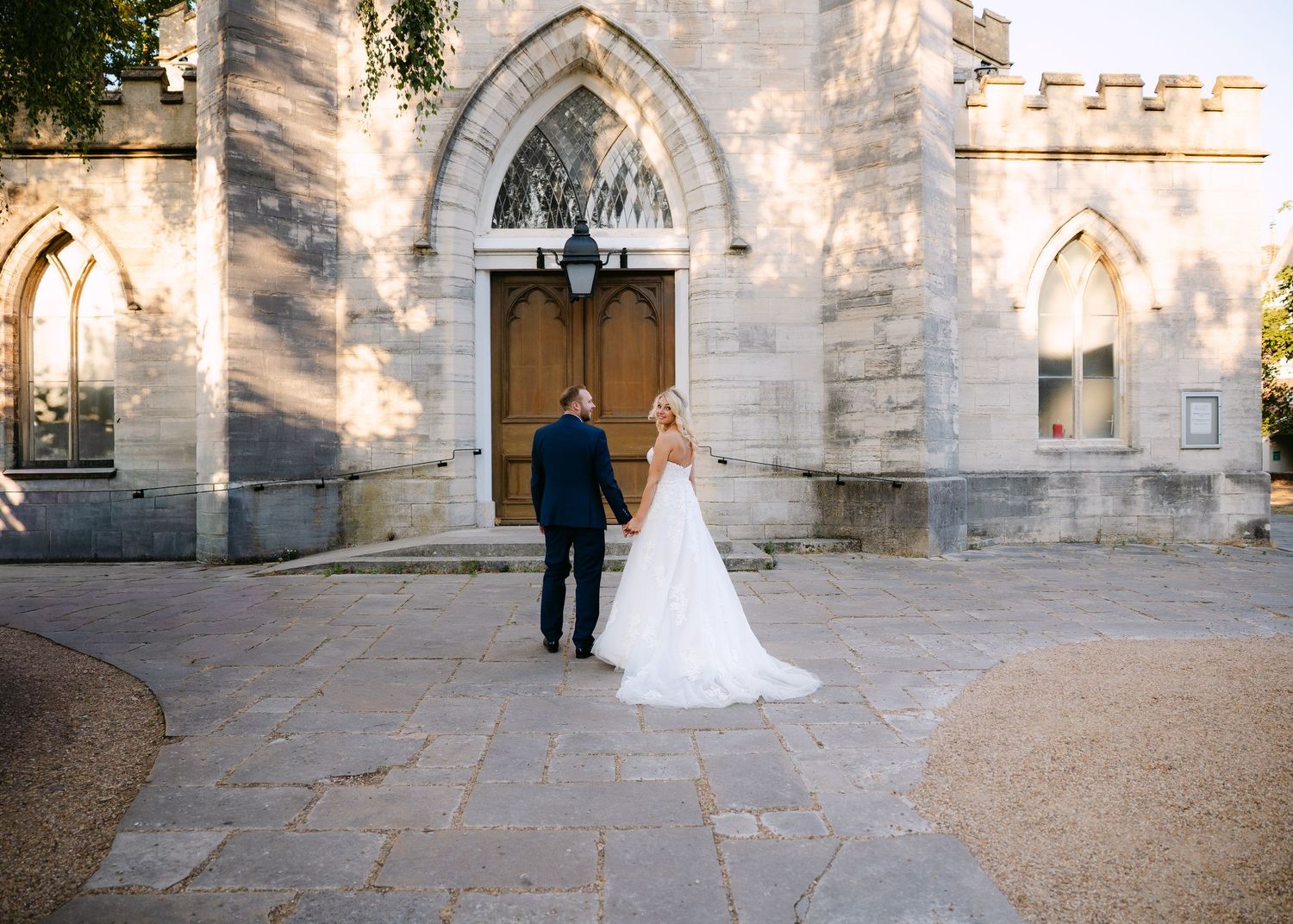 Bride and groom stand hand in hand before a historic stone church with Gothic arched doors on their wedding day.