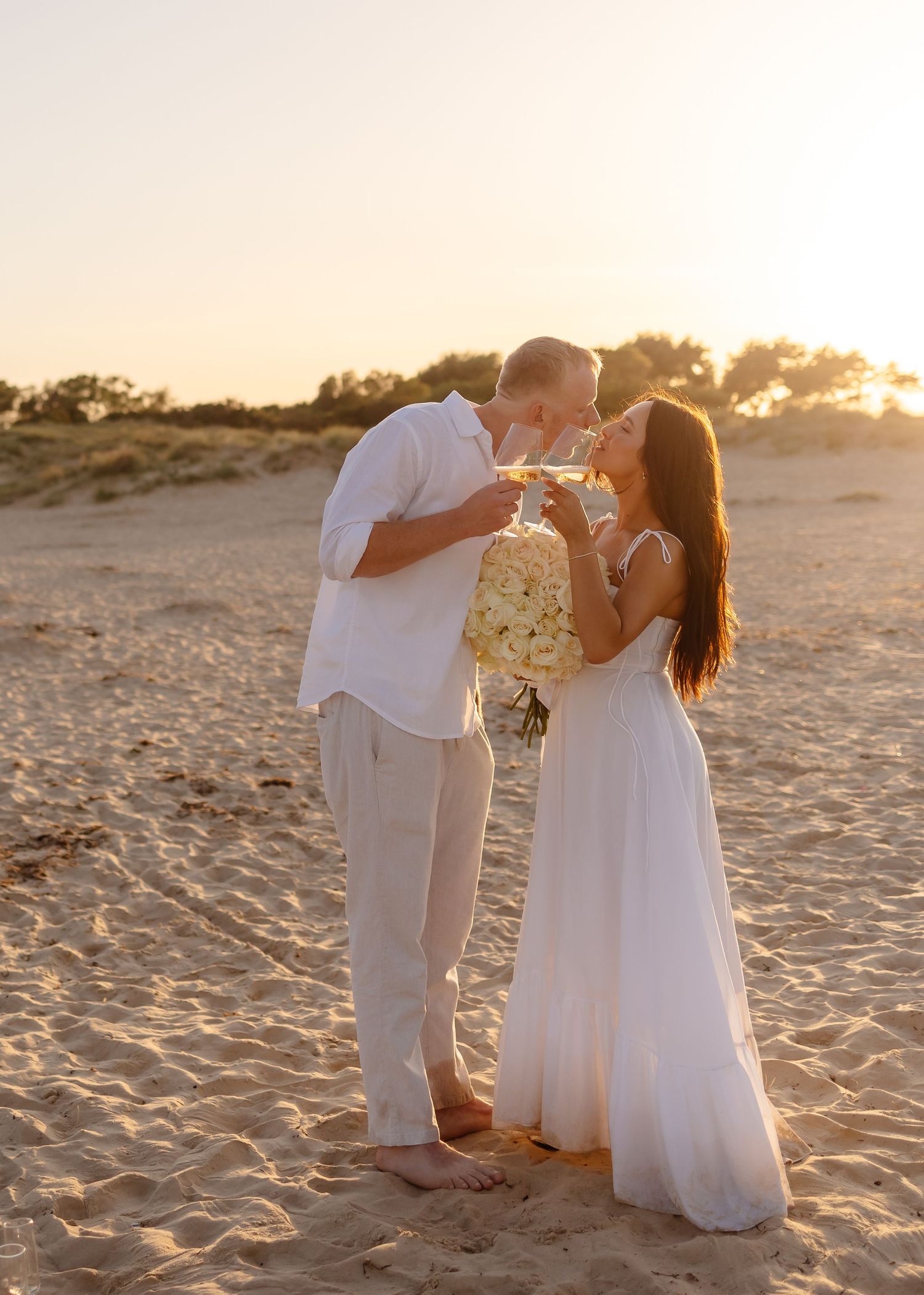 Couple in white attire sharing a kiss at golden sunset on sandy beach, woman holding white floral bouquet.
