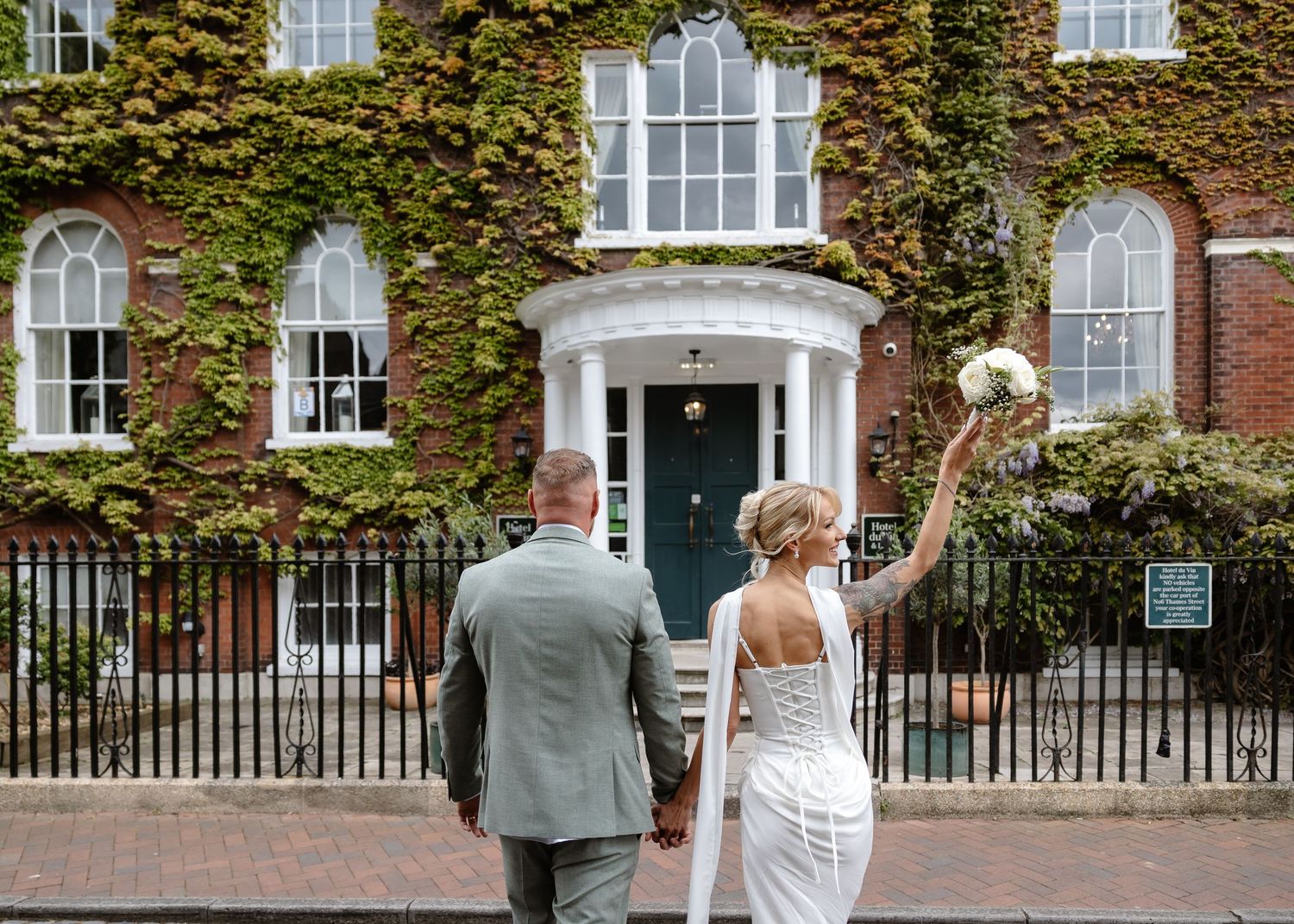Bride and groom holding hands outside ivy-covered brick wedding venue with teal door, bride raising bouquet.