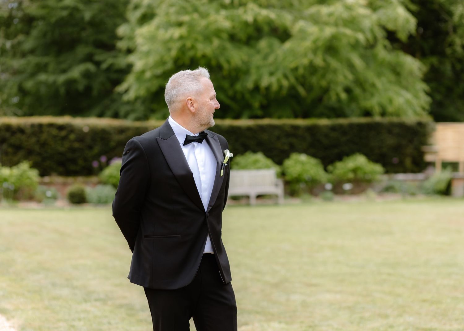 Elegant groom in black tuxedo and bow tie standing outdoors in lush garden setting on wedding day.