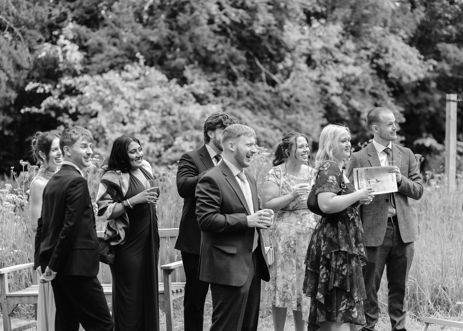 Black and white photo of elegantly dressed wedding guests gathered outdoors, socializing and holding programs.
