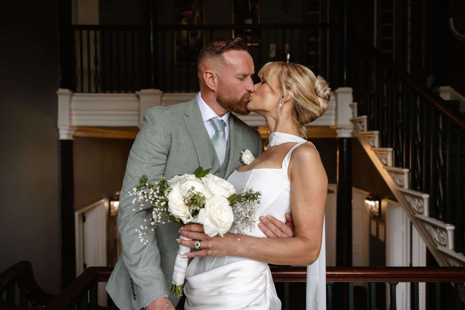 Bride and groom share a romantic kiss indoors, bride holding white floral bouquet, groom in light gray suit.