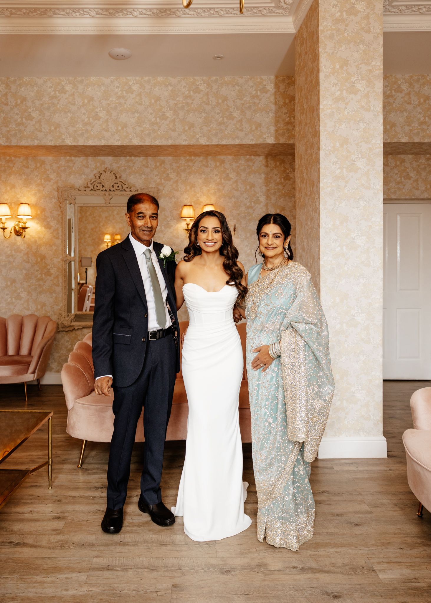 A wedding celebration in an elegant room with floral wallpaper, featuring three people in formal attire including a sari.