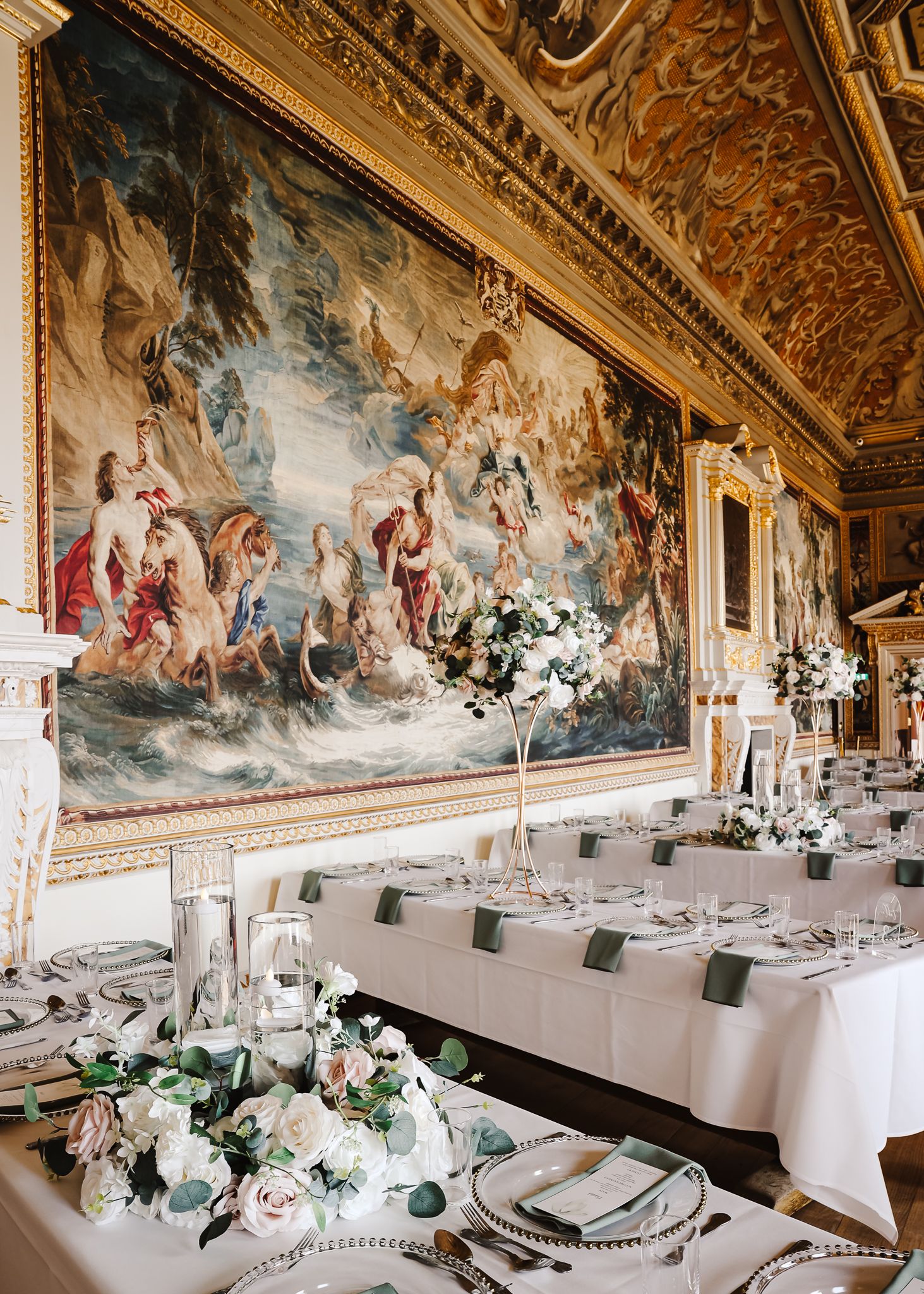 Elegant banquet hall with ornate ceiling murals, white floral centerpieces and formal table settings in a palatial room.