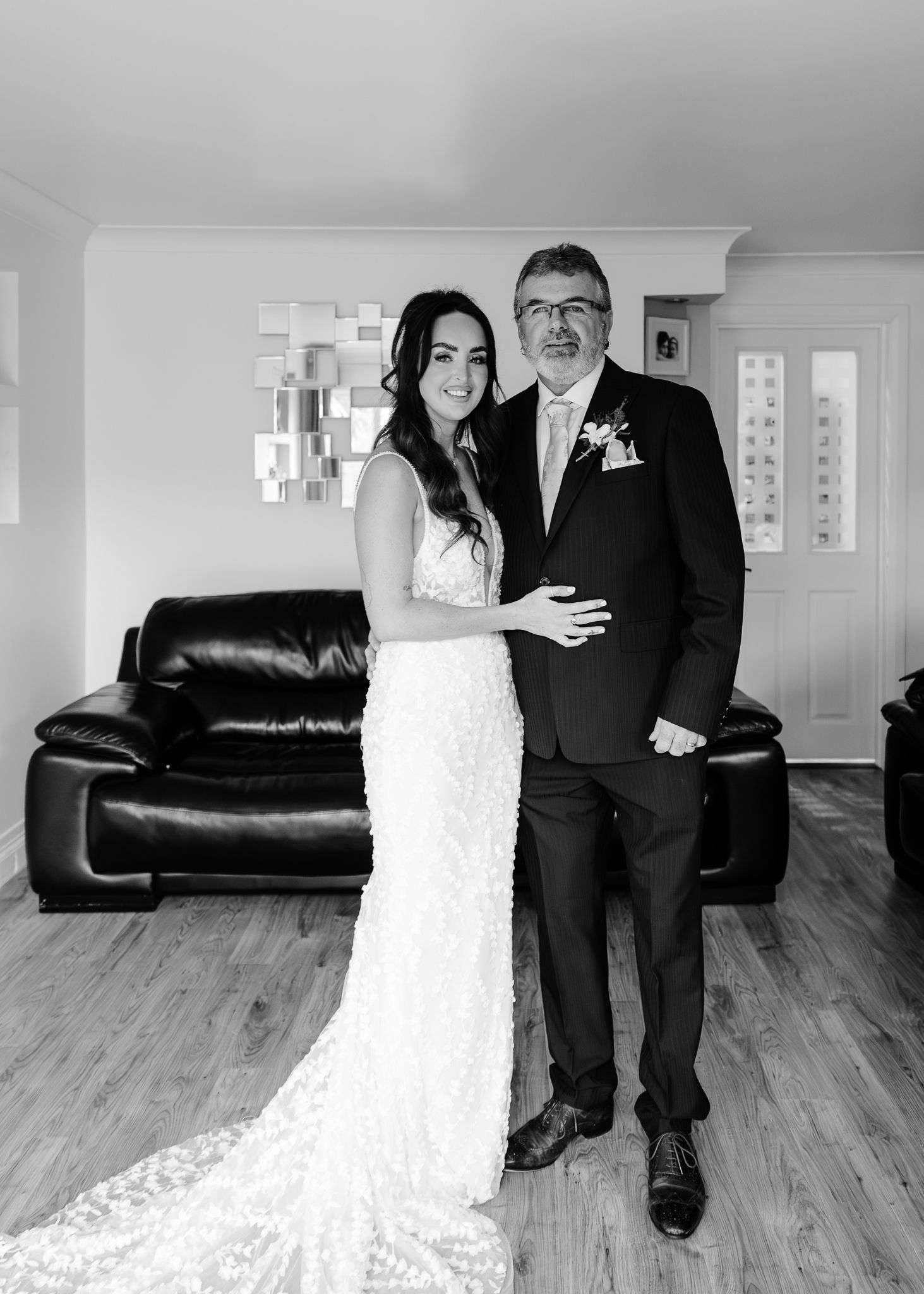 A couple in wedding attire sits together on a black leather couch in a bright room with wooden floors.