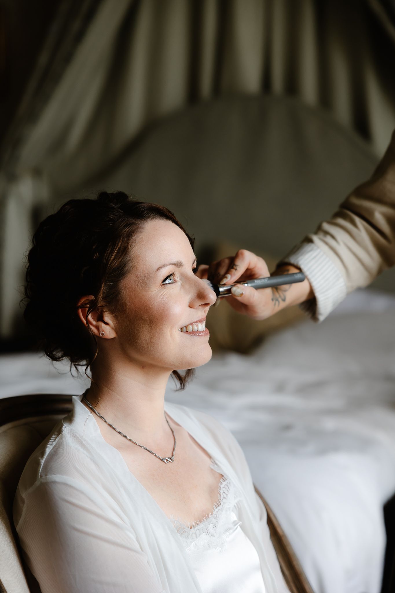 A person having makeup applied while wearing a white robe in a softly lit room.