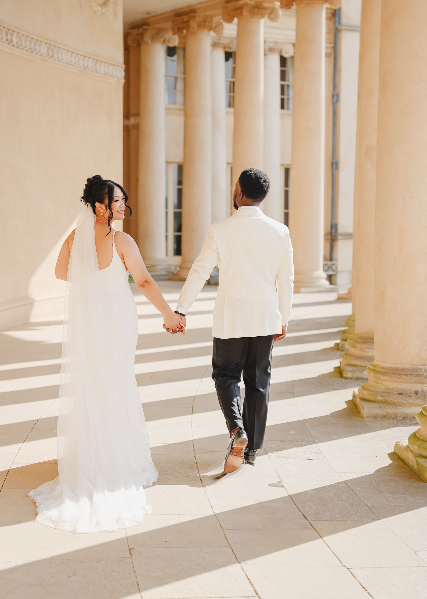 Couple walks hand in hand through sunlit colonnade with classical architecture.