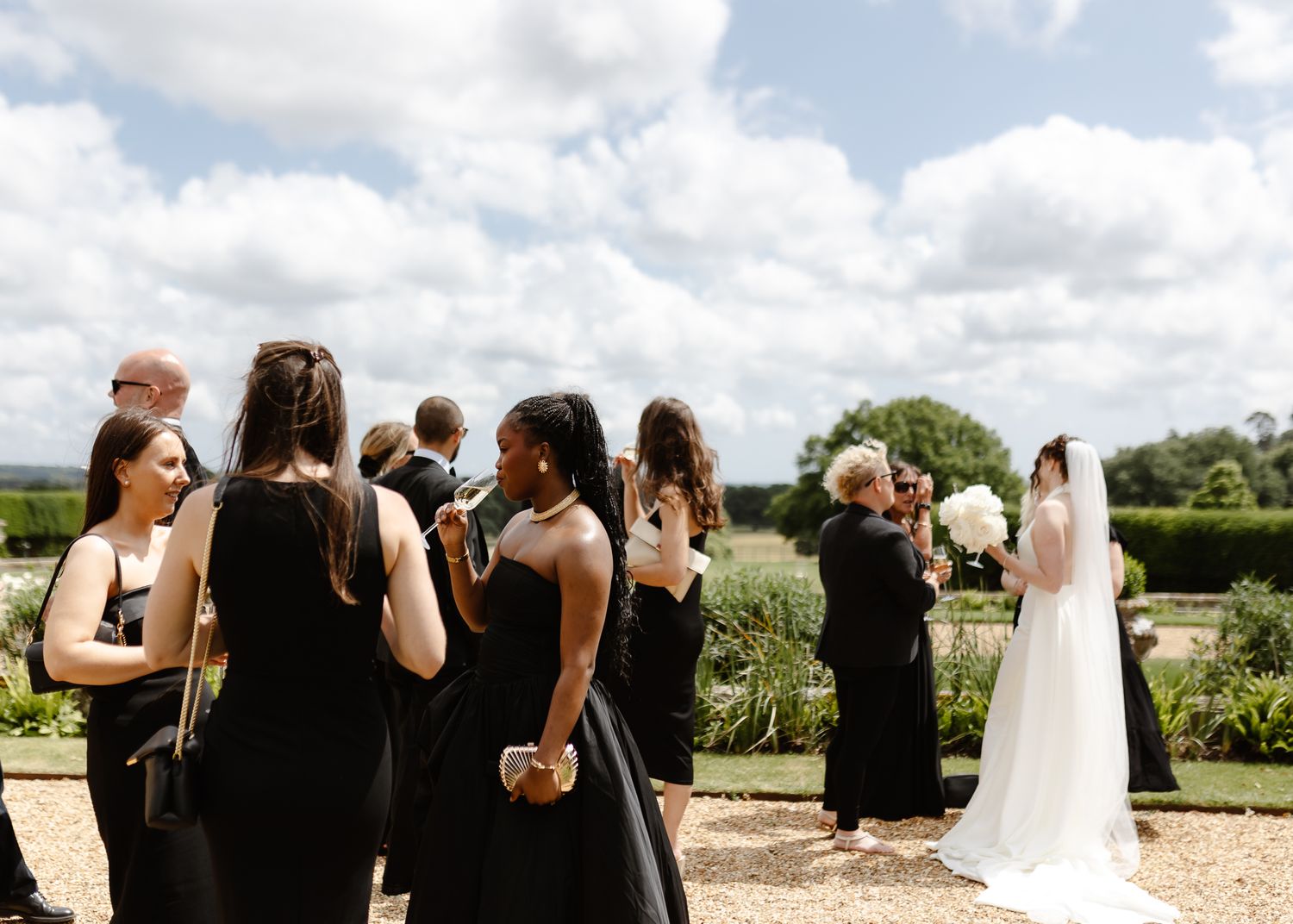 A panoramic view of a wedding ceremony gathering outdoors with guests mingling on a gravel path surrounded by greenery.
