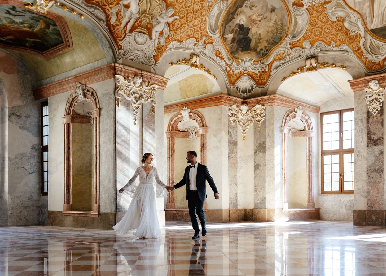 Panoramic view of newlyweds dancing in a sunlit baroque palace hall with golden frescoes and marble columns.