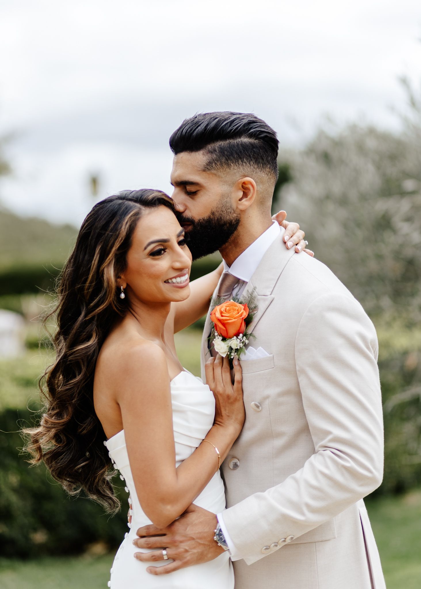 A romantic wedding portrait shows a couple embracing outdoors, with the groom in a cream suit and coral boutonniere.