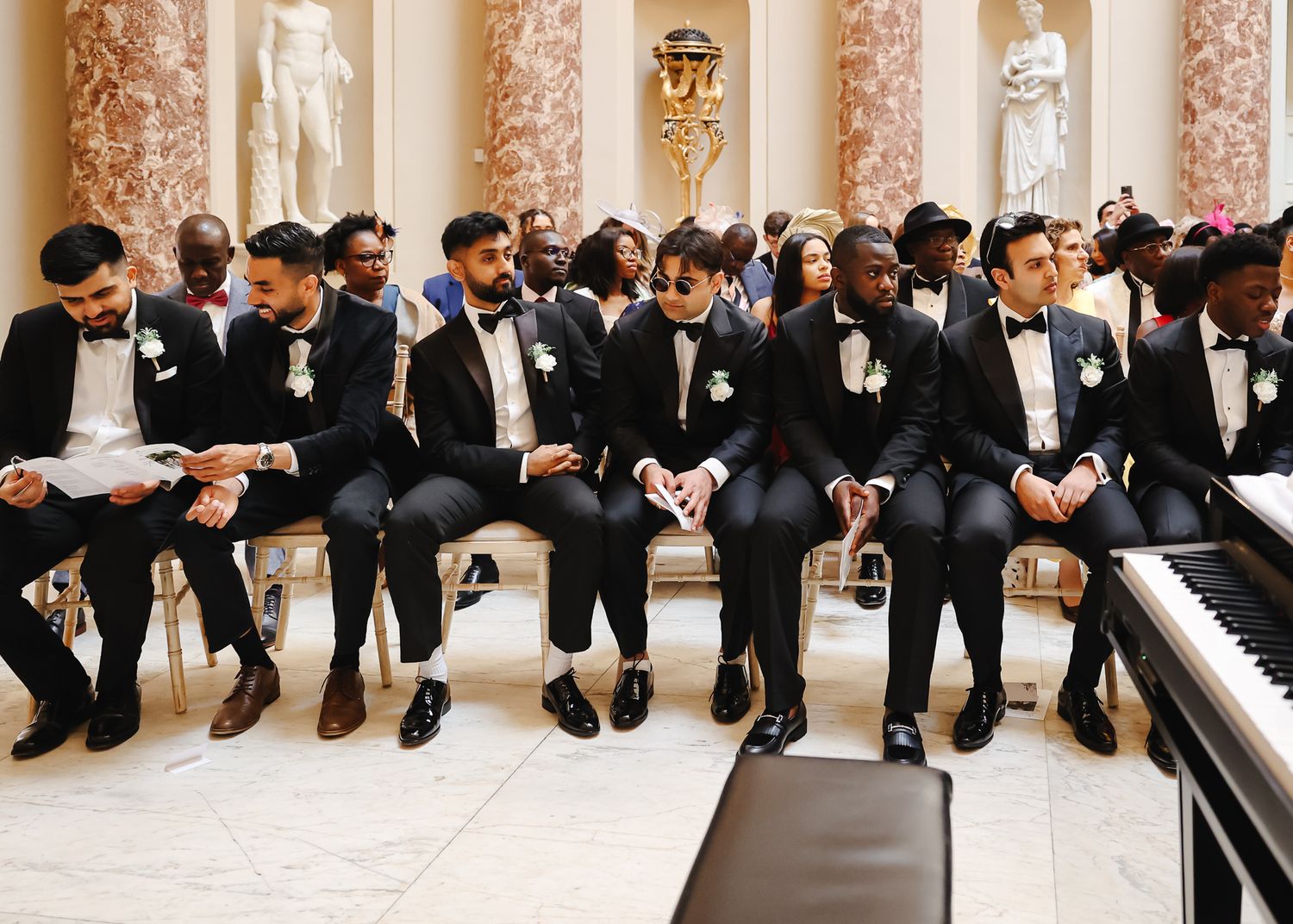Groomsmen in black tuxedos sit together on a bench in a formal venue with marble columns.