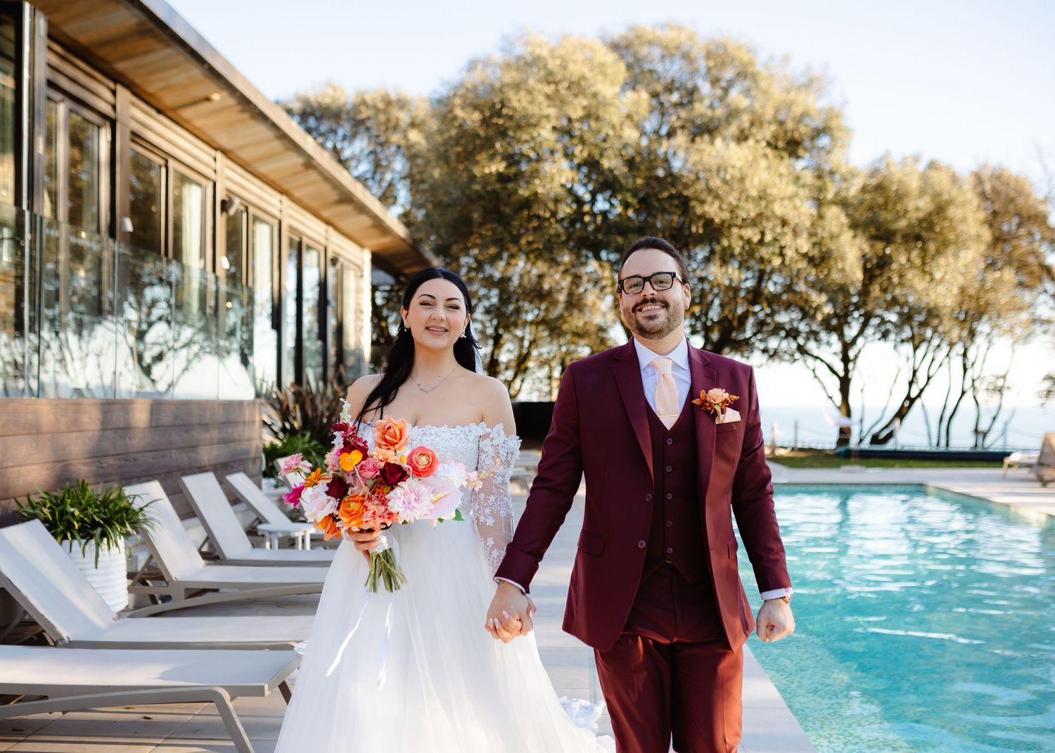 Bride and groom holding hands by a pool, she in a white gown with bouquet, he in a burgundy suit.