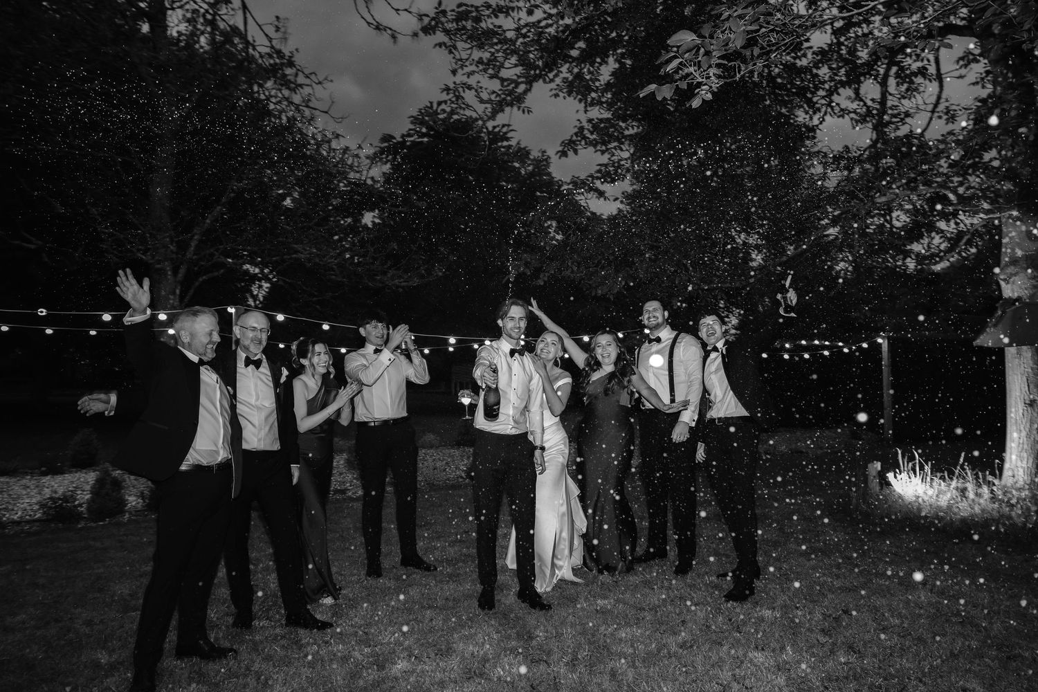 Black and white wedding photo of bride, groom, and guests celebrating outdoors at night with string lights.