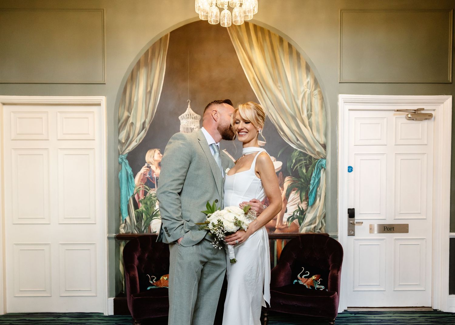 Bride and groom share a romantic kiss in an elegant venue with ornate mural backdrop and chandelier on their wedding day.