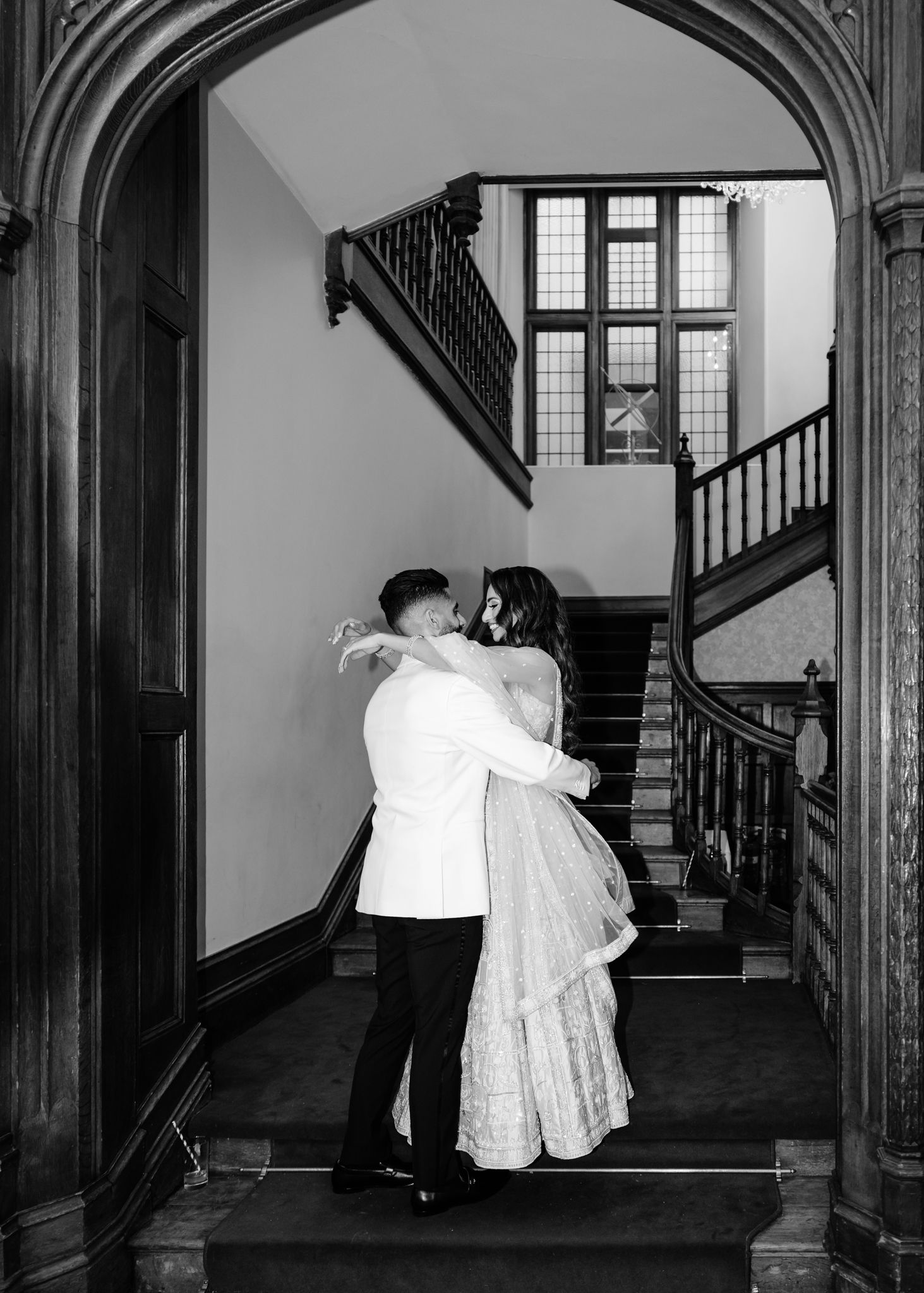 A romantic couple embraces on an elegant staircase with gothic arched doorway and vintage architectural details.
