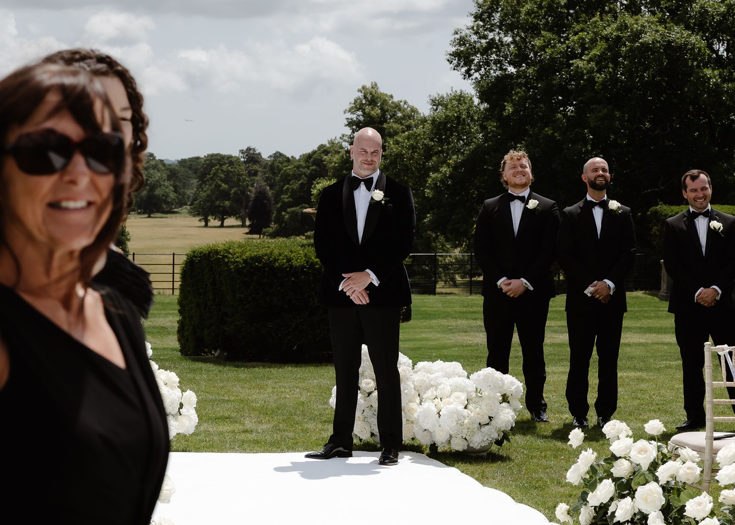 A wedding party poses outdoors on a grassy lawn with white floral arrangements and trees in the background.