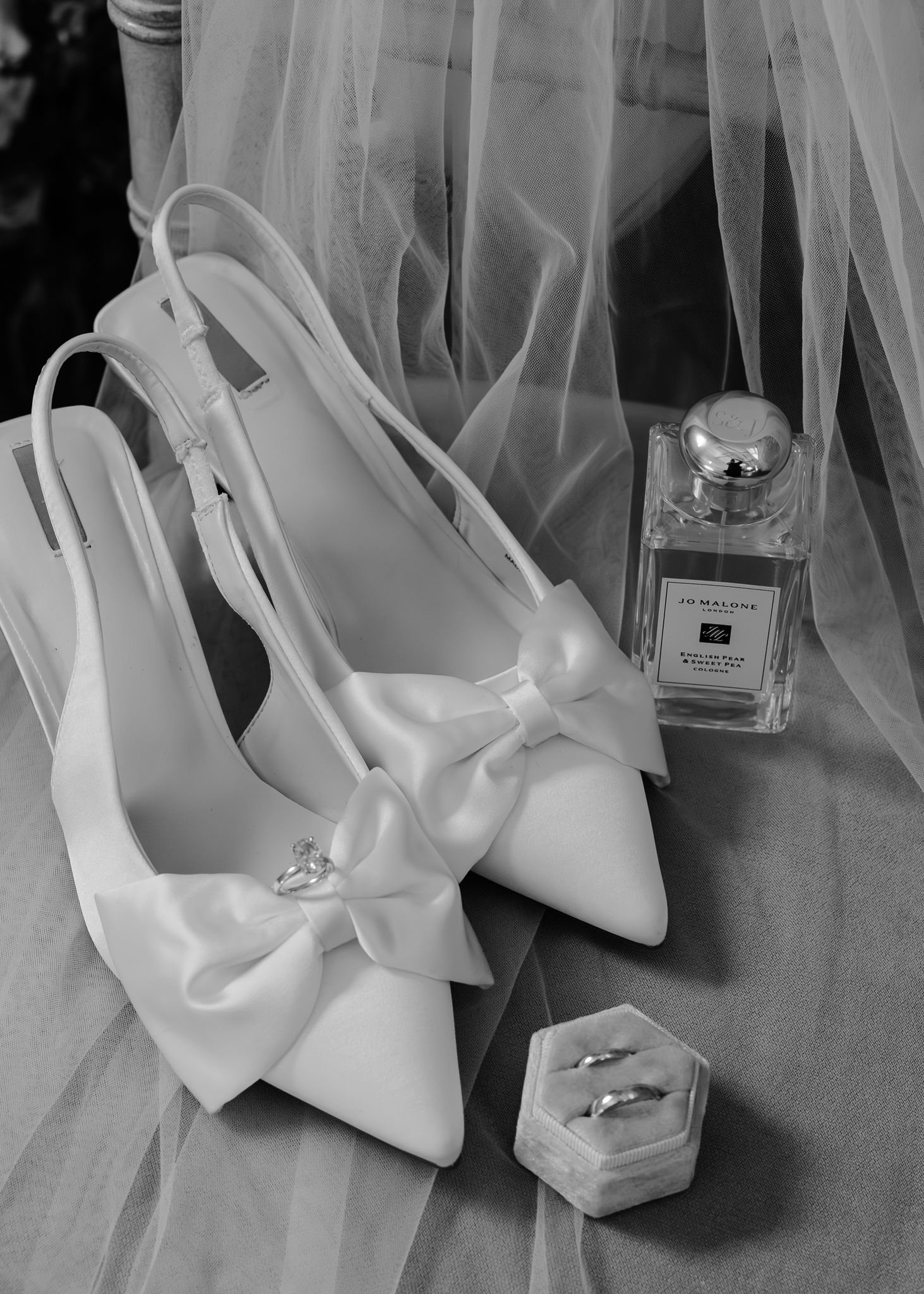 White bridal heels with elegant bows displayed alongside perfume bottles and wedding accessories in a monochromatic setting.