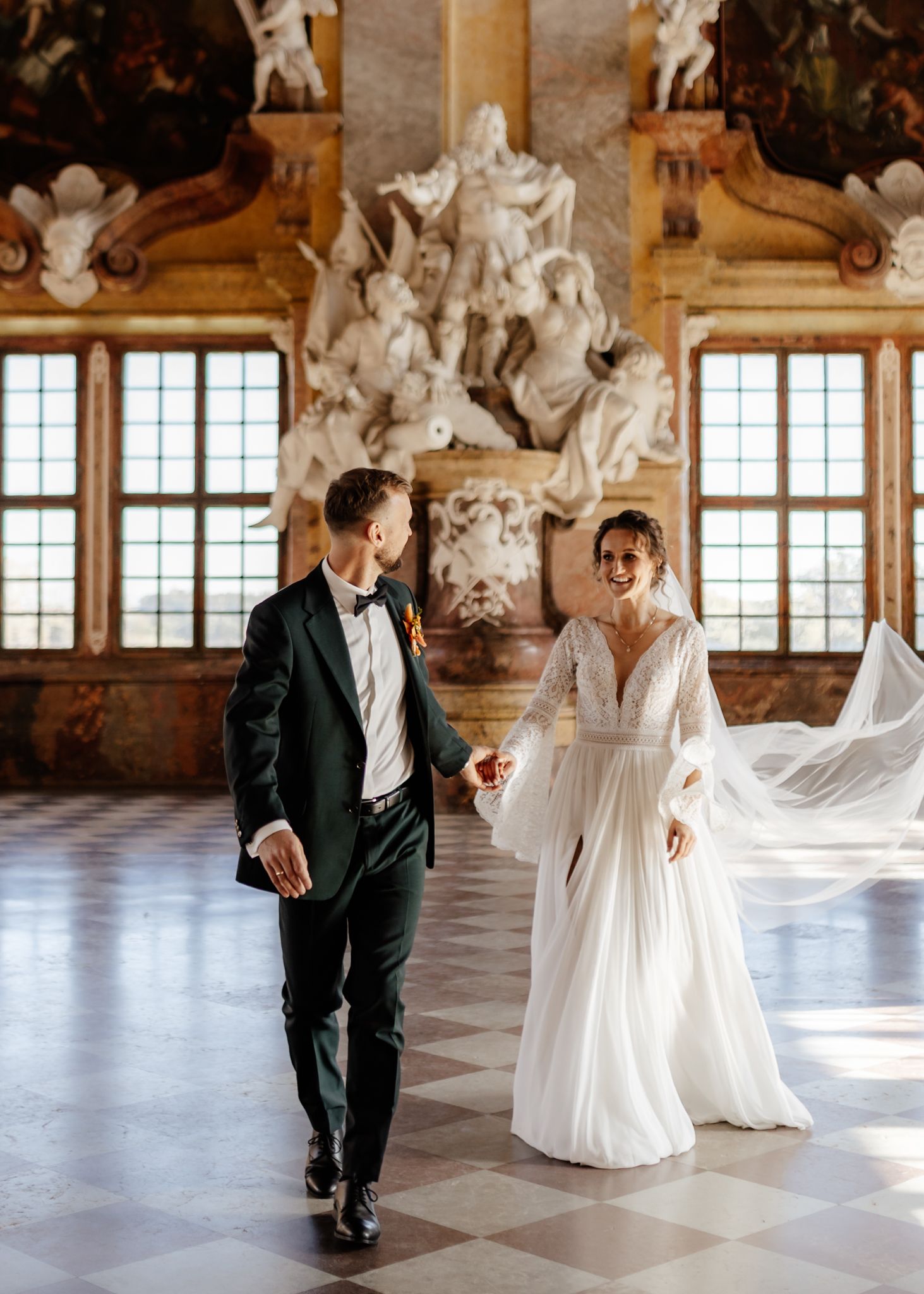 A couple dances gracefully in a grand baroque ballroom with ornate white sculptures and large windows.