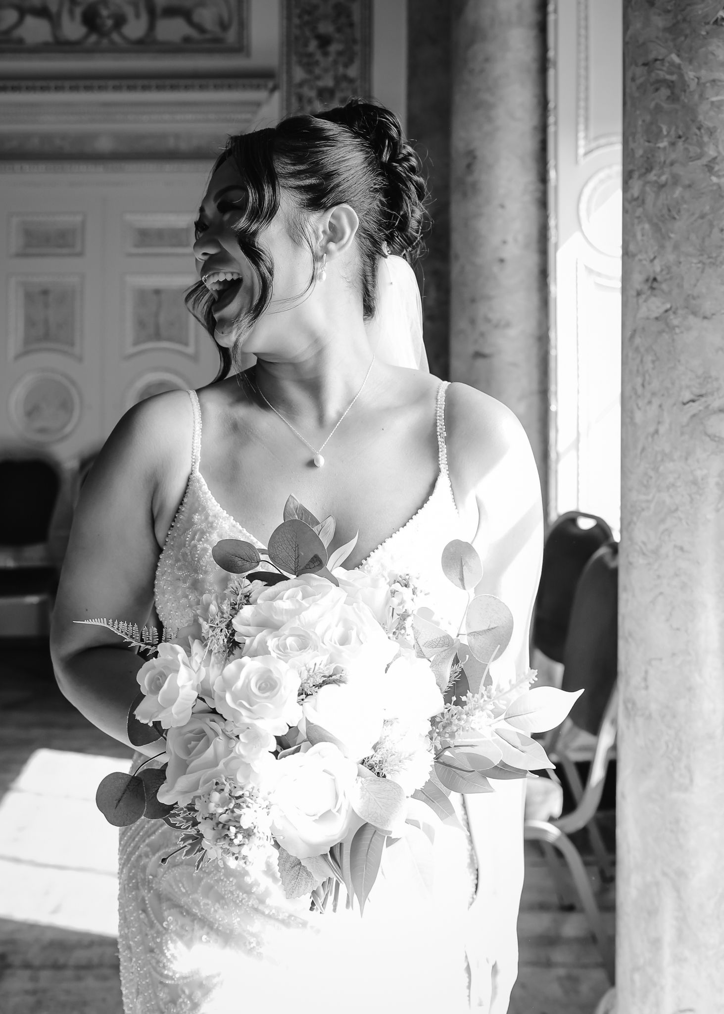 A bride in a white dress laughs joyfully while holding a bouquet of white flowers in an elegant venue.