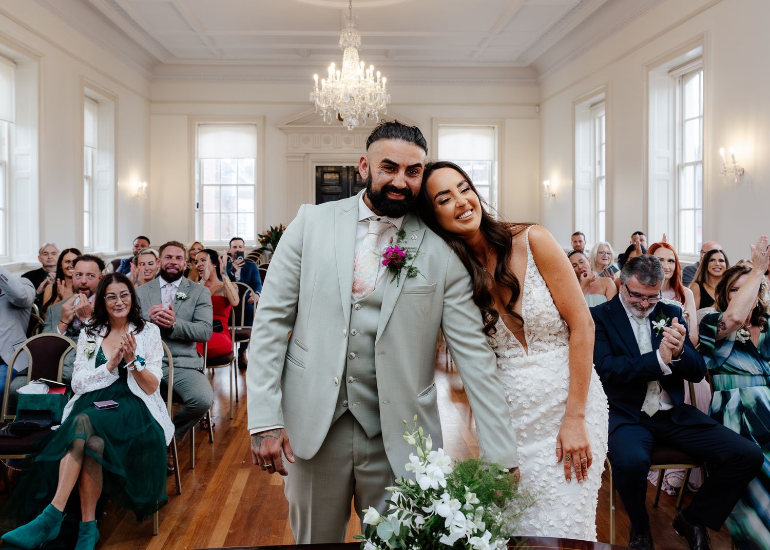 Elegant wedding ceremony in bright white room with crystal chandelier and guests seated in rows.