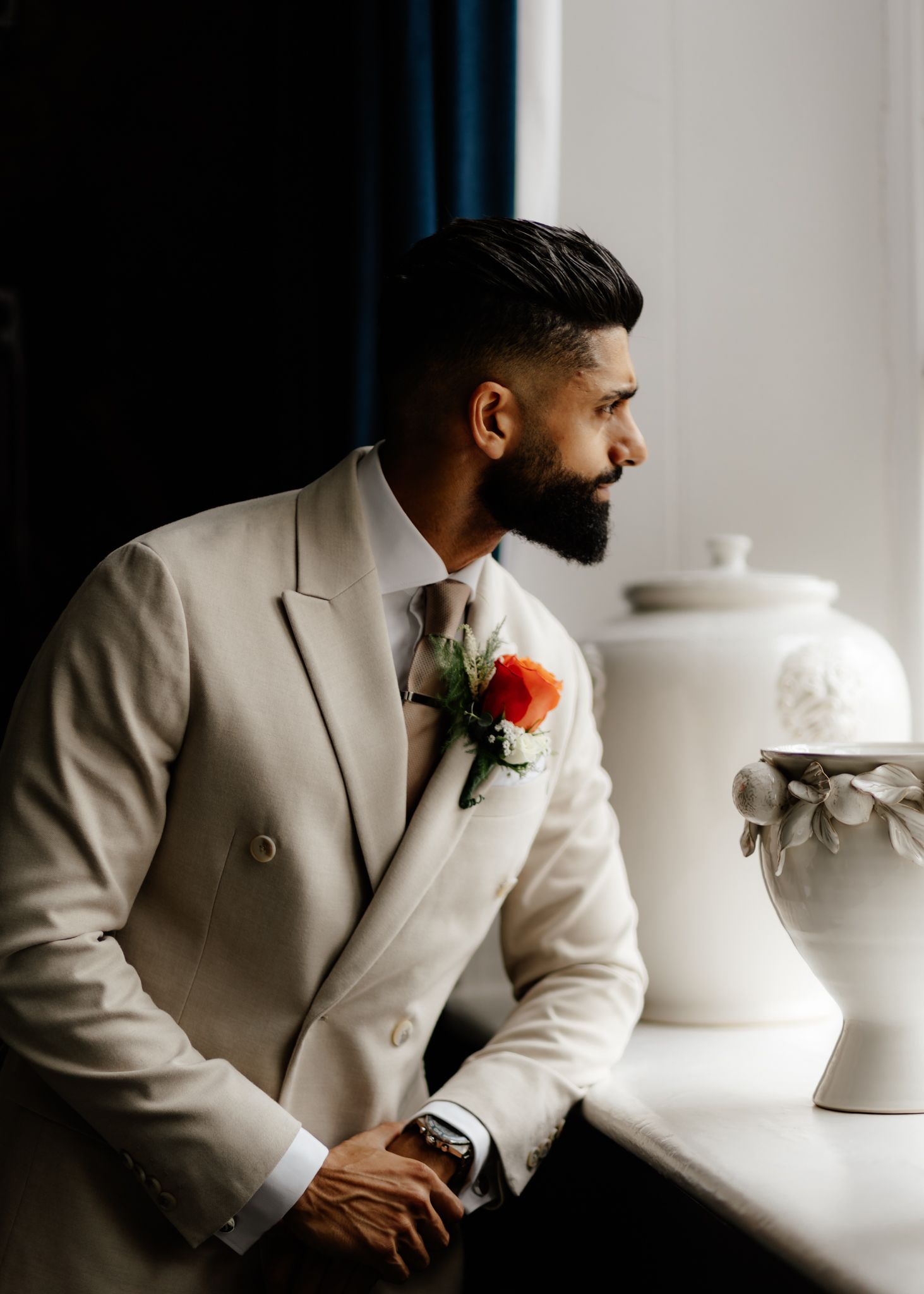 A groom in a beige double-breasted suit with a red boutonniere gazes thoughtfully by a white vase in dramatic lighting.