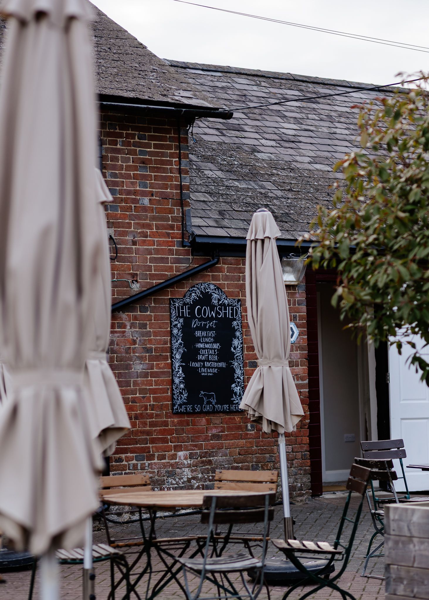 Outdoor cafe terrace with folded beige umbrellas and black chairs against a rustic brick building facade.