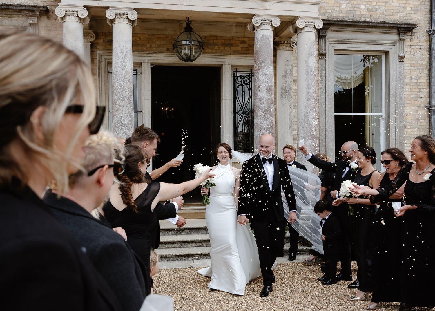 Wedding guests throw confetti at a couple on the steps of an elegant stone building with classical columns.