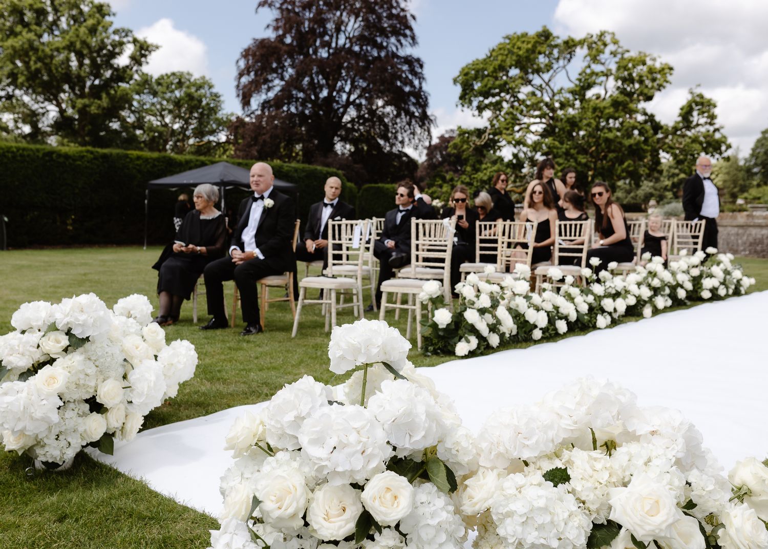 Outdoor wedding ceremony setup with white flowers lining the aisle and chiavari chairs on the lawn.