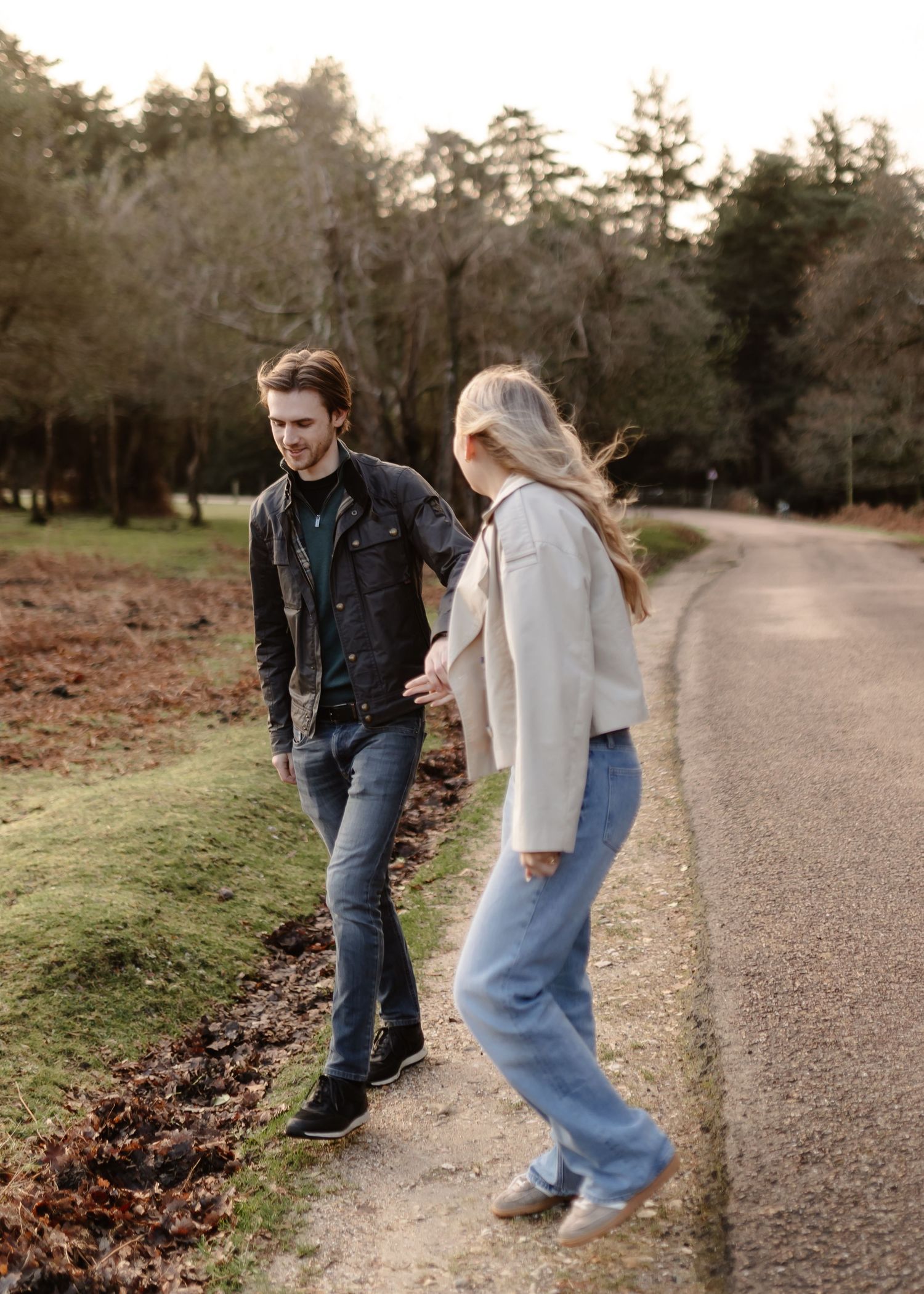 A young couple walks along a tree-lined path, smiling at each other on a cool autumn day.