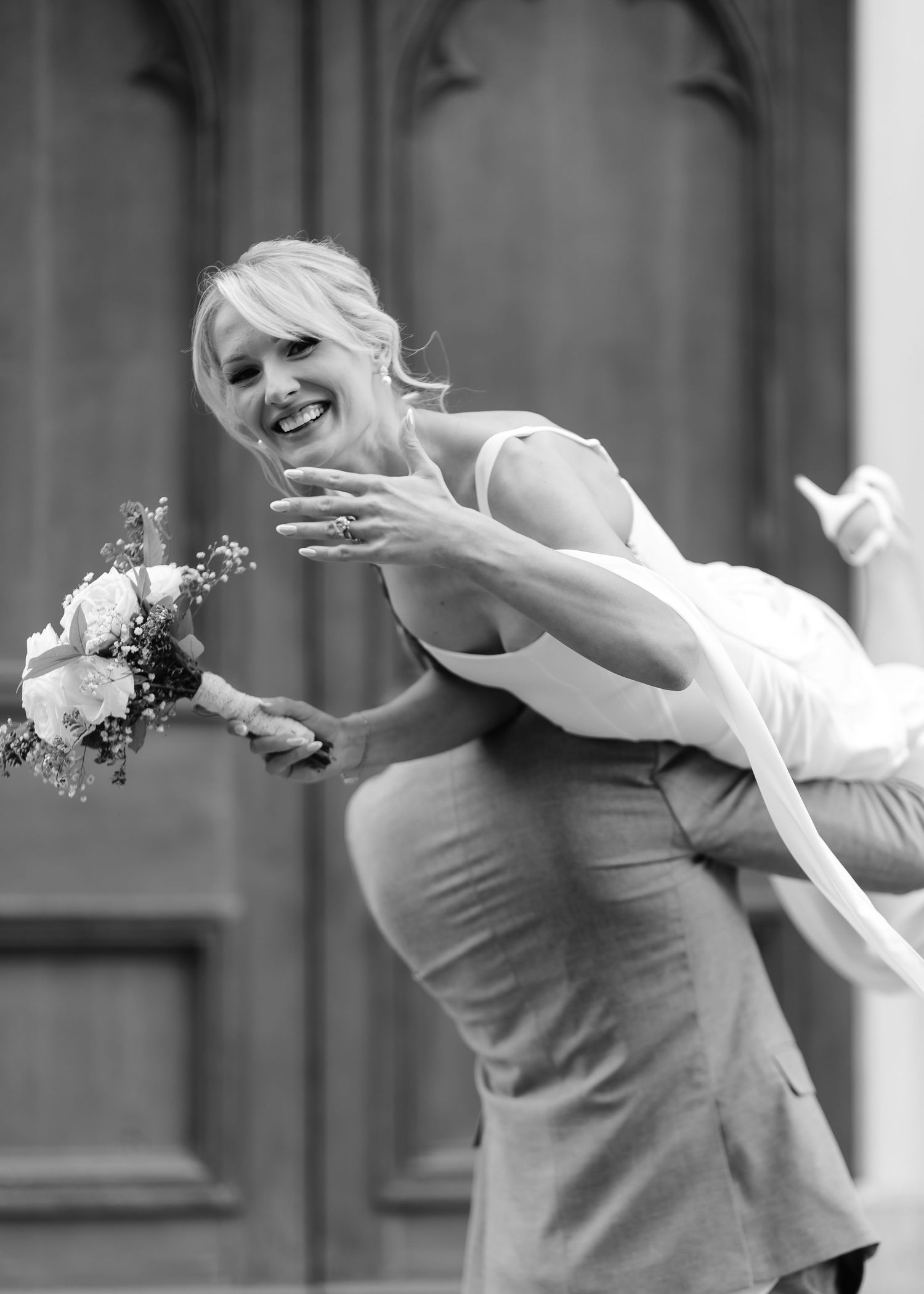 Black and white photo of a smiling bride being lifted by groom, holding a bouquet outside a church.