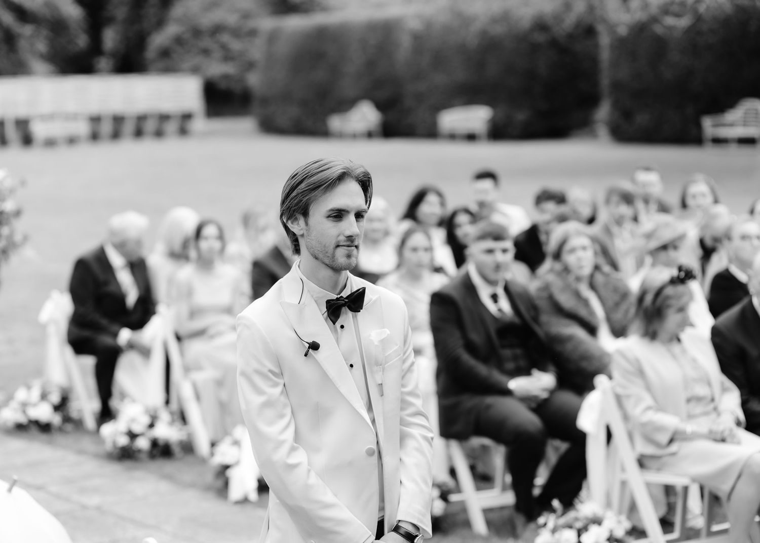 Groom in white tuxedo standing at outdoor wedding ceremony aisle with seated guests in background, black and white photo.