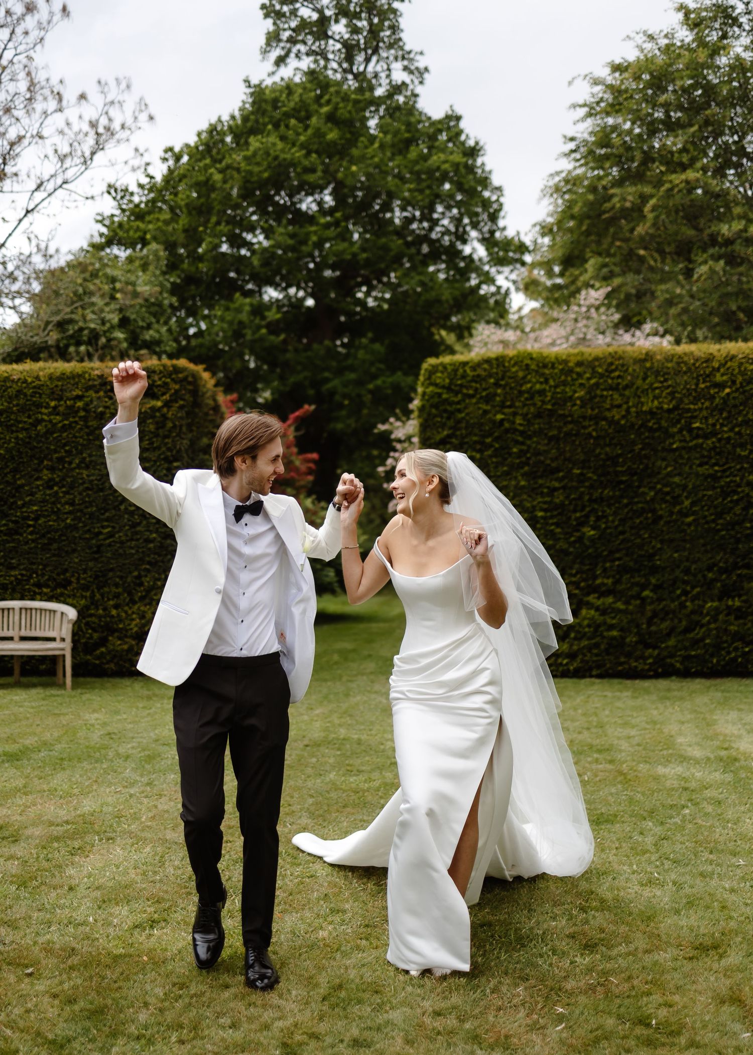 Joyful bride and groom celebrating in garden, bride in satin gown with veil, groom in white tuxedo jacket.