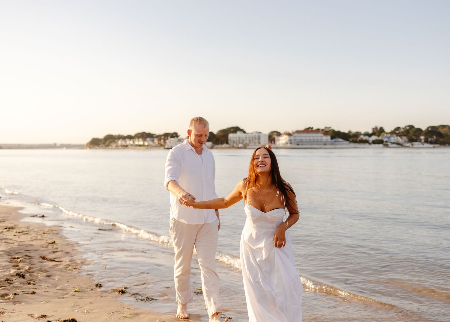 Couple walking hand in hand on sunny beach at golden hour, woman in white dress.
