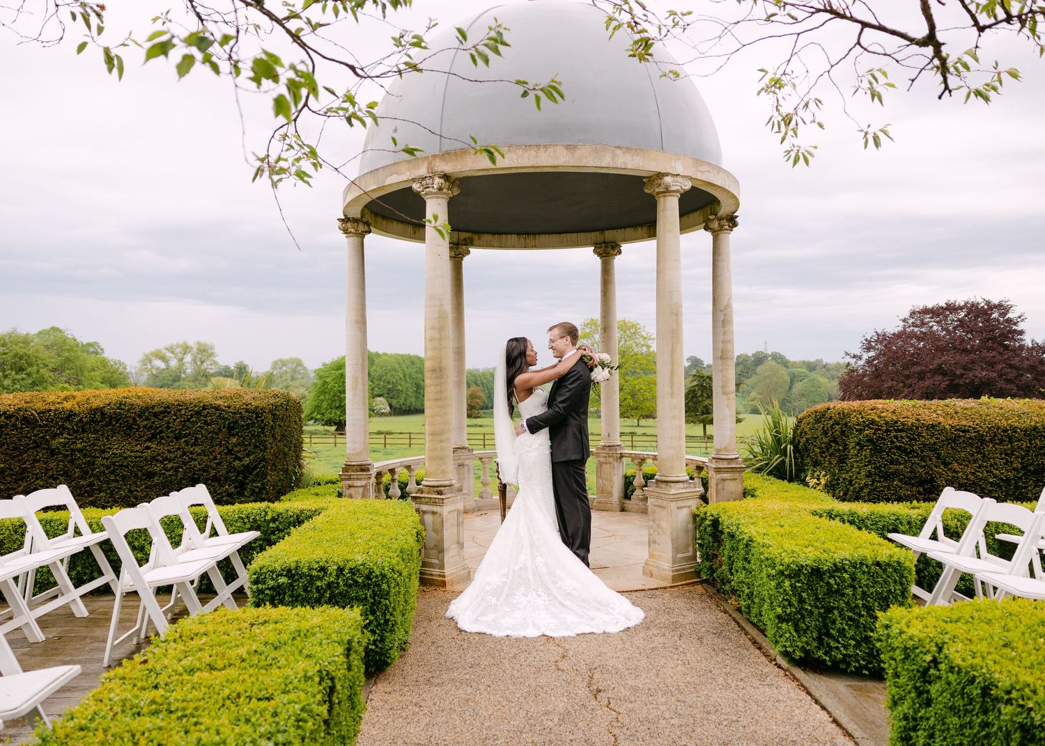 Bride and groom share a kiss under an elegant stone gazebo surrounded by manicured hedges and white chairs at an outdoor wedding.