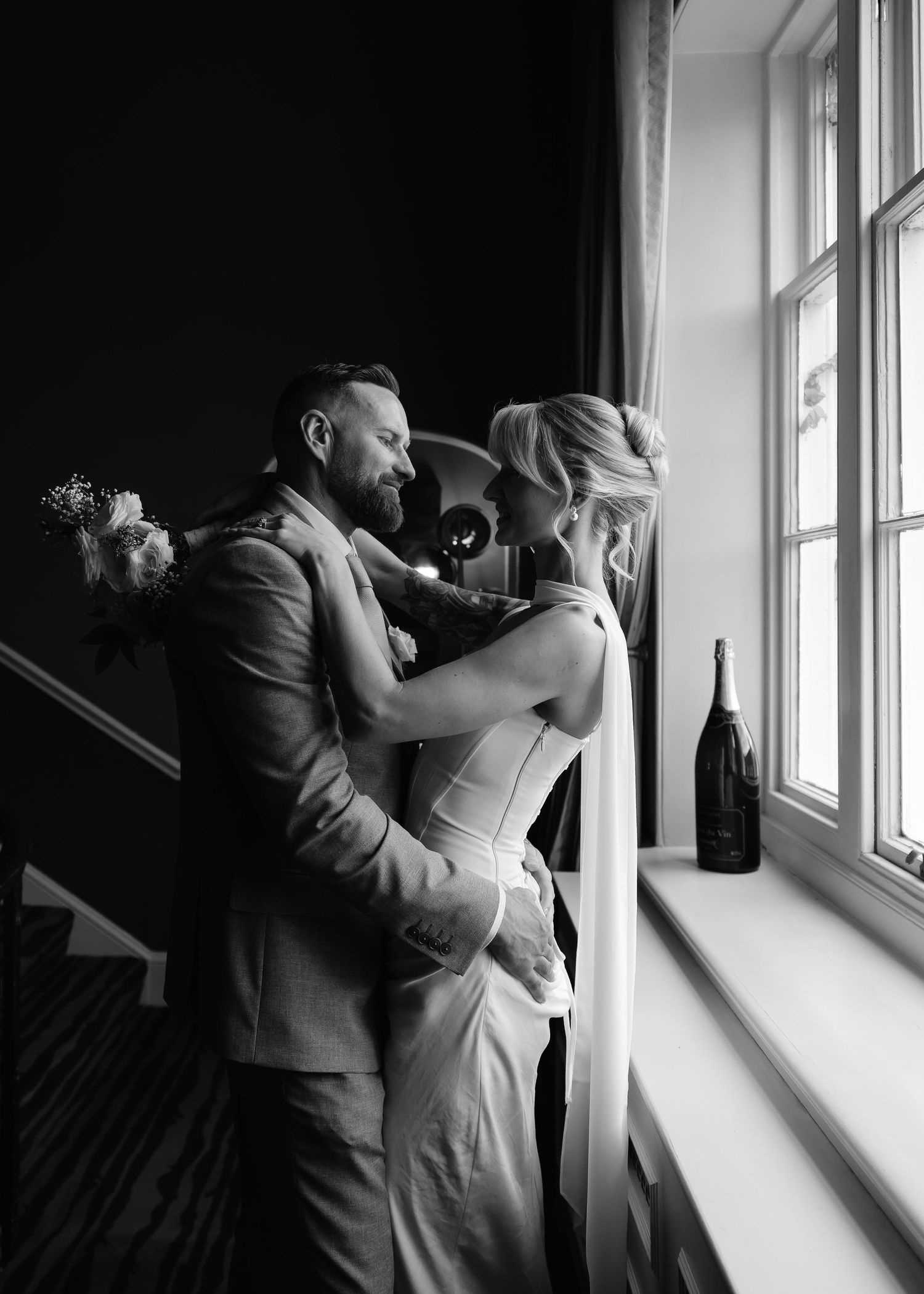 Black and white photo of bride and groom embracing romantically by a window with champagne bottle nearby.