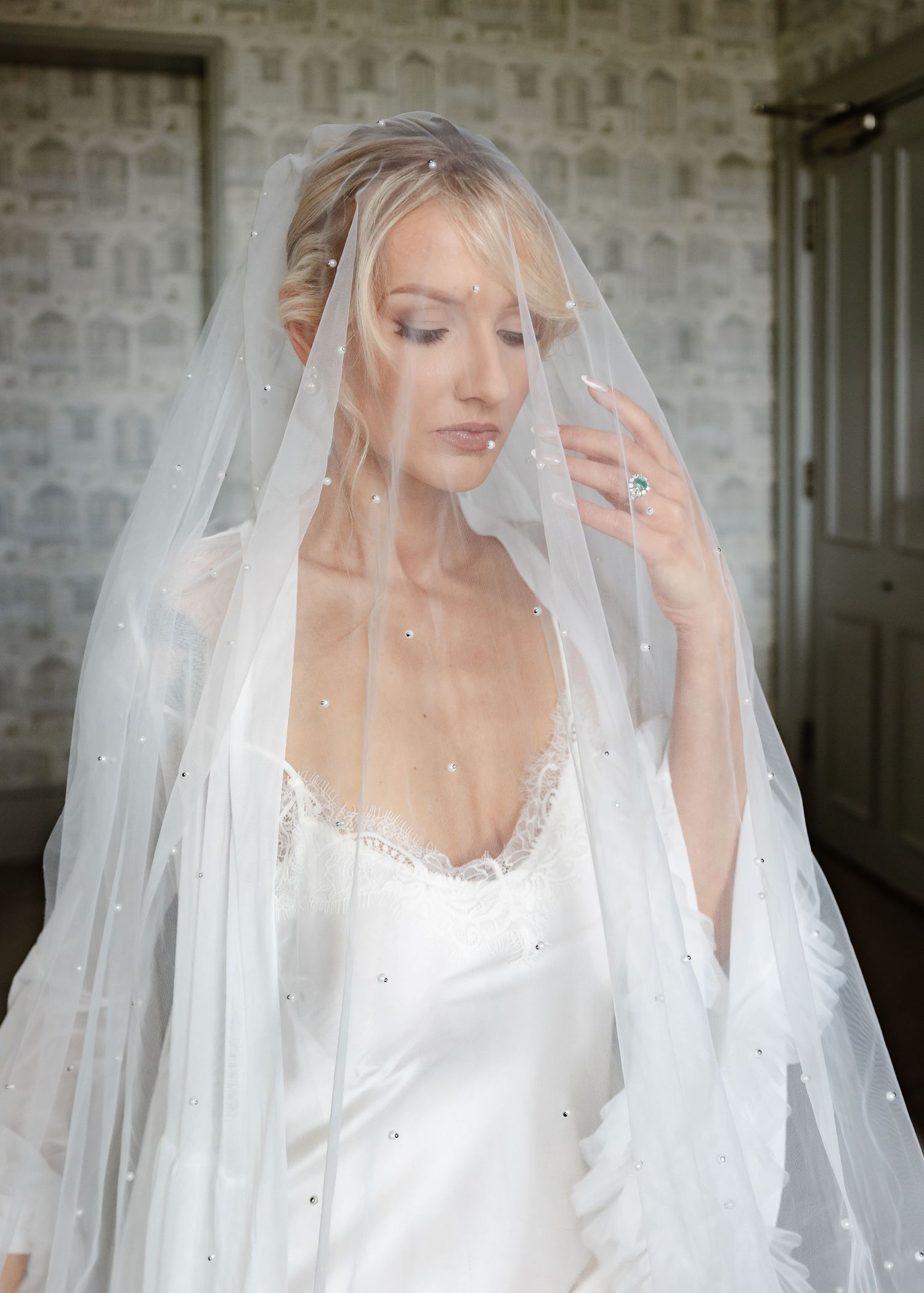 Bride wearing a pearl-embellished wedding veil and satin gown, posing elegantly in a softly lit room.