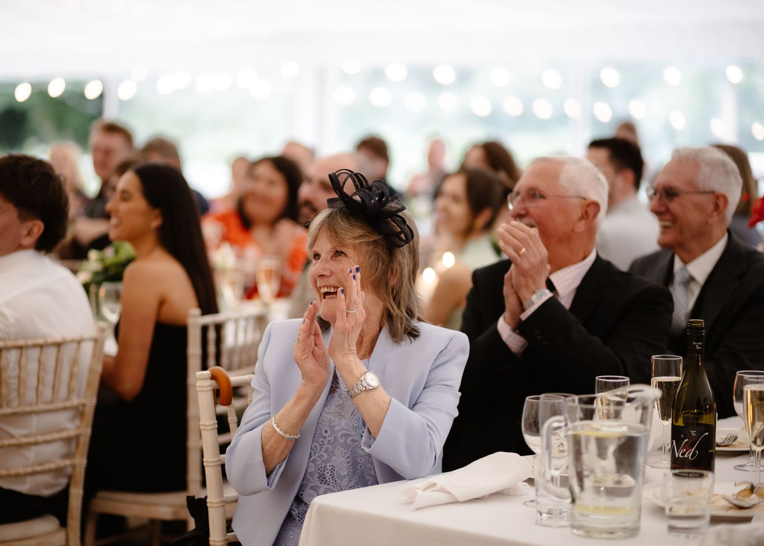 Wedding guests clapping and smiling at a reception in a marquee, woman in blue jacket wearing a fascinator.