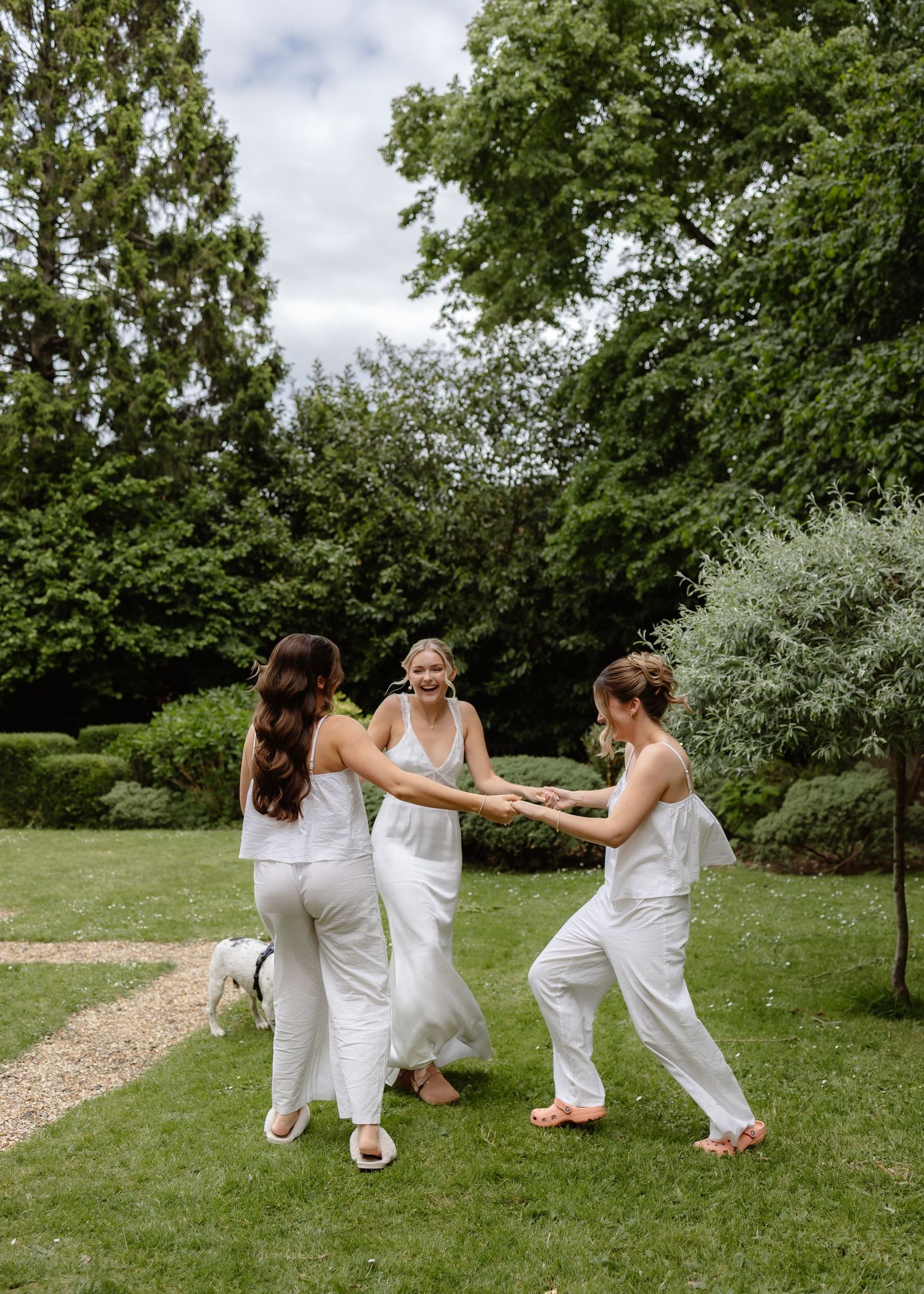 Bride in white gown laughs with two bridesmaids in white outfits while holding hands on a lush green garden lawn.