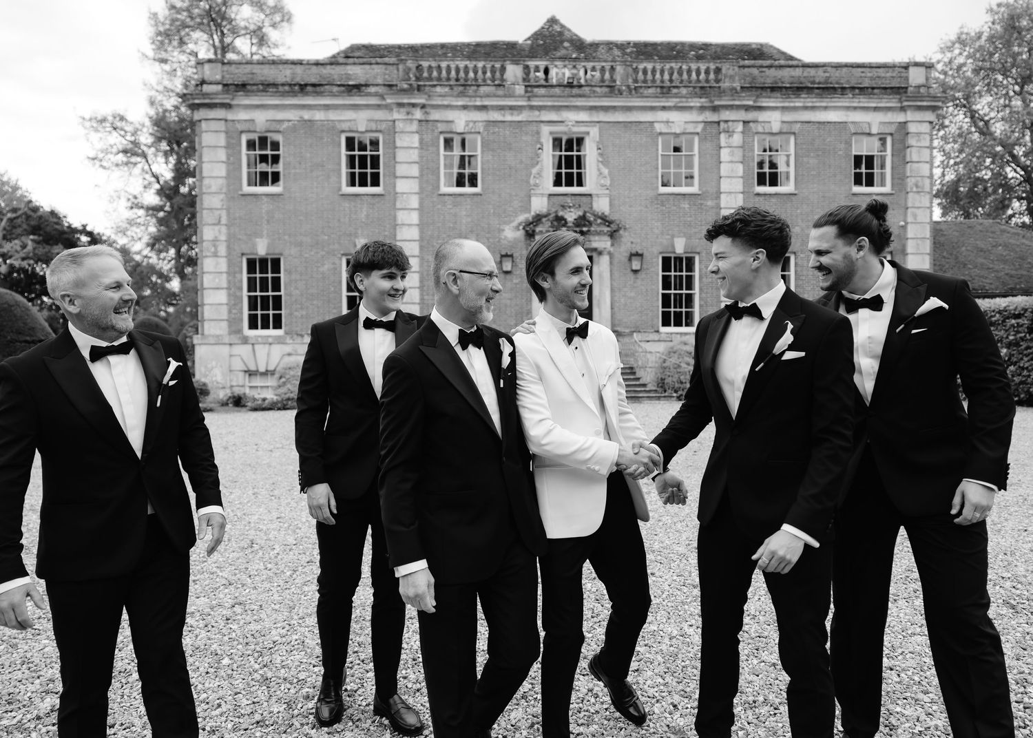 Black and white photo of groom in white tuxedo shaking hands with groomsmen in black suits outside a manor house.